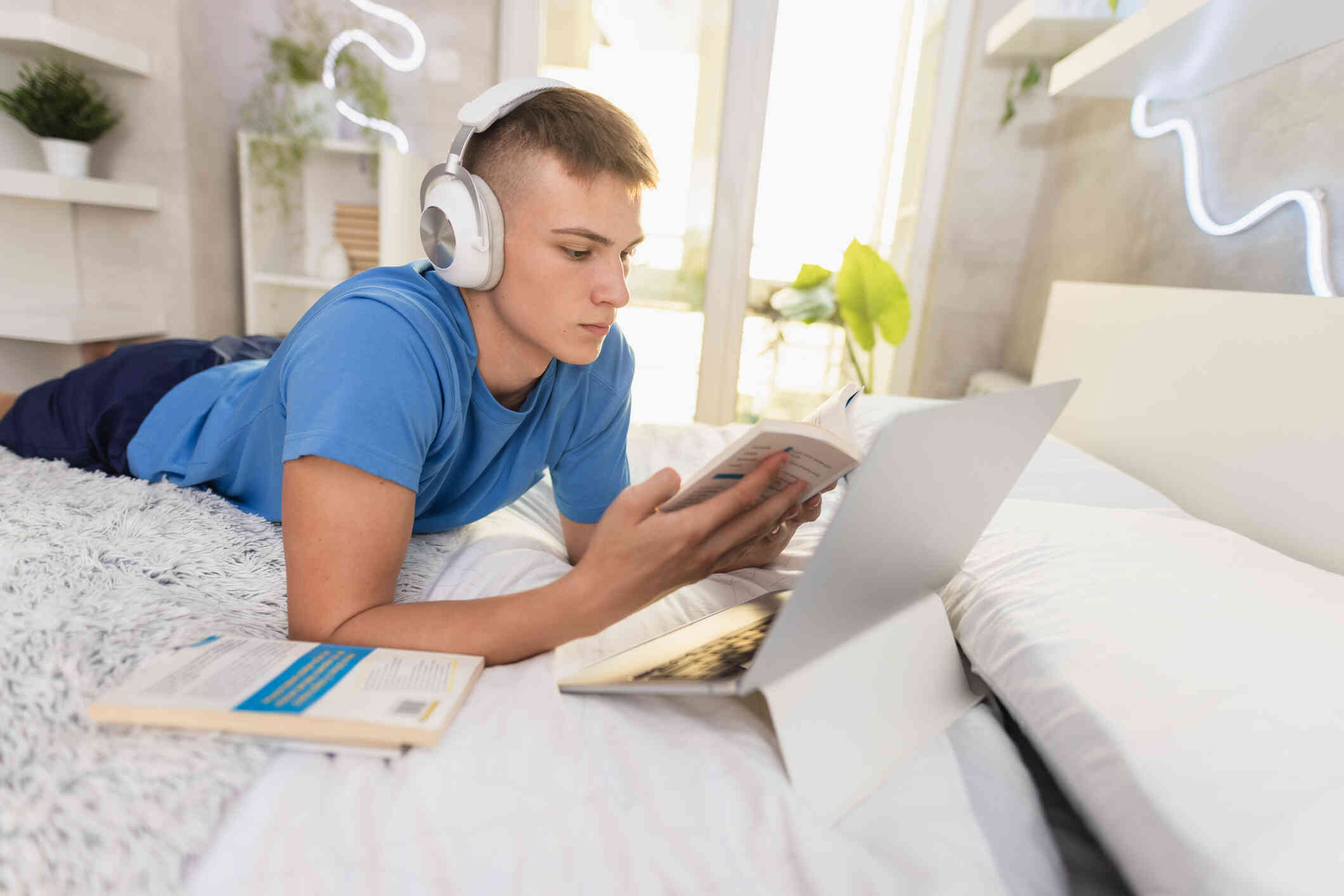 A teen boy in a blue shirt lays on his stomach on the bed while wearing headphones with his laptop open infront of him as he reads from a book in his hands.