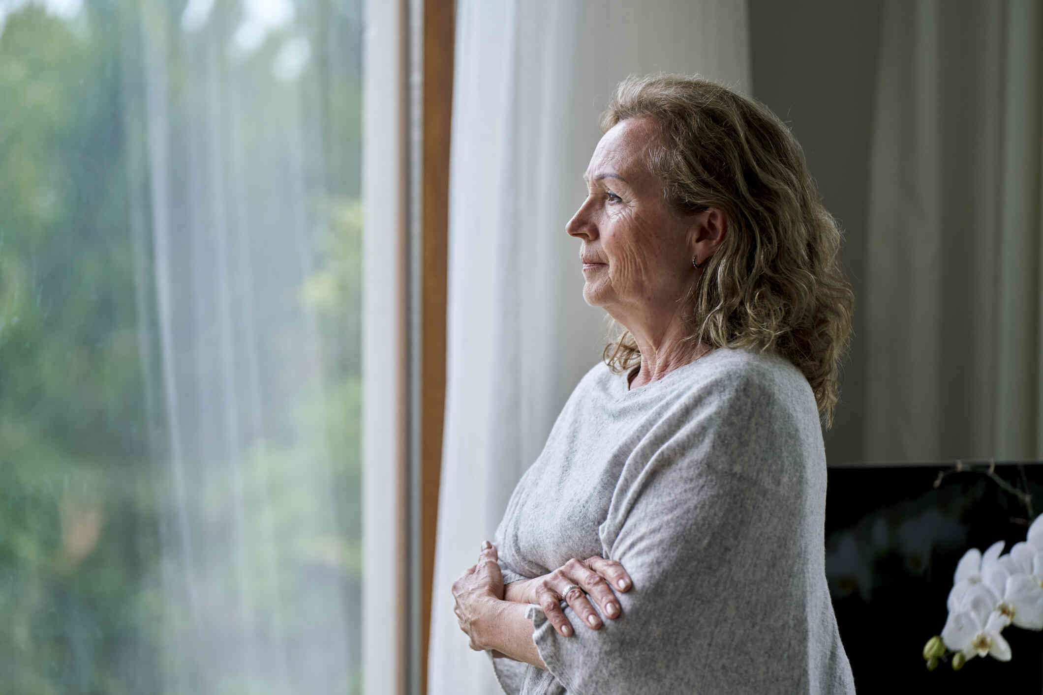 A middle aged woman in a grey sweater crosses her arms while sadly gazing out of the window of her home.