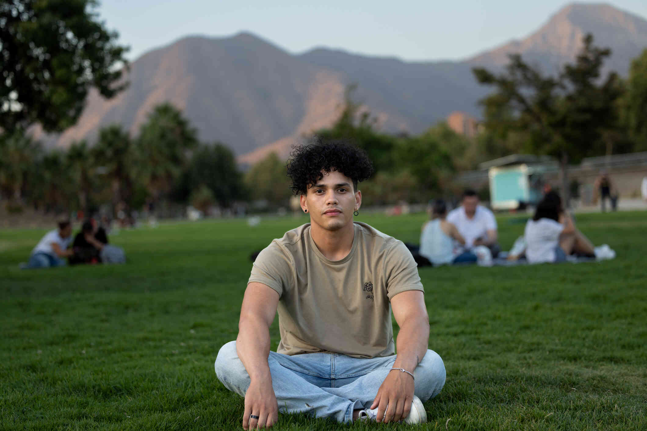 A man in a tab shirt sits cross-legged on the grass in a courtyard  and looks at the camera with a serious expression.