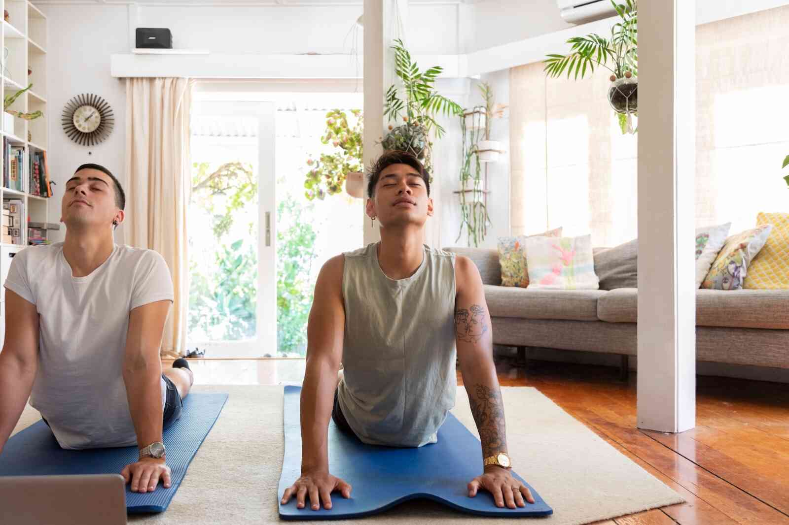 Two men lay on yoga mats while doing yoga poses and looking at a laptop screen. 