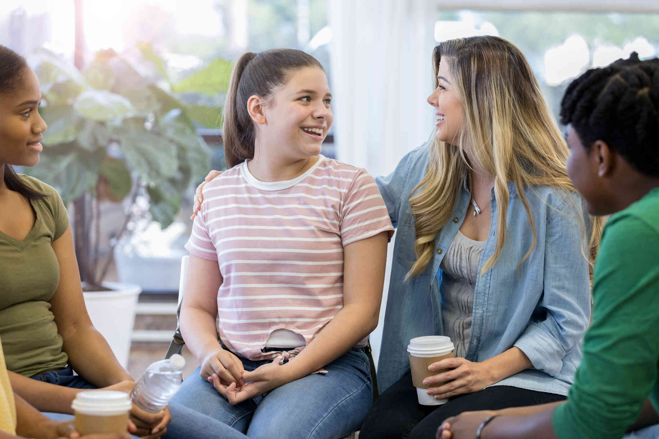 A teenage girl in a pink striped shirt smiles at a friend who has her arm around her as they sit with a group of other young people holding coffee cups.