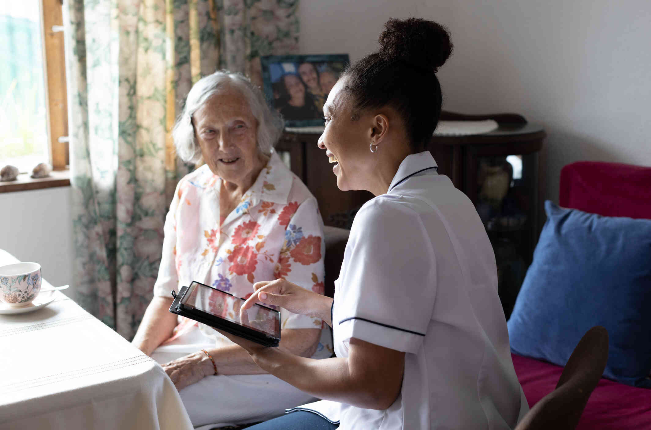 A female nurse sits next to her female elderly patient as they smile at one another during in home care.