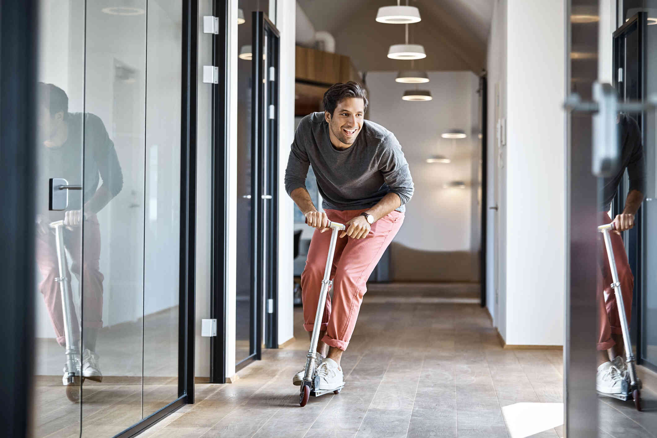 A man rolls down a hallways on a scooter while smiling brightly.