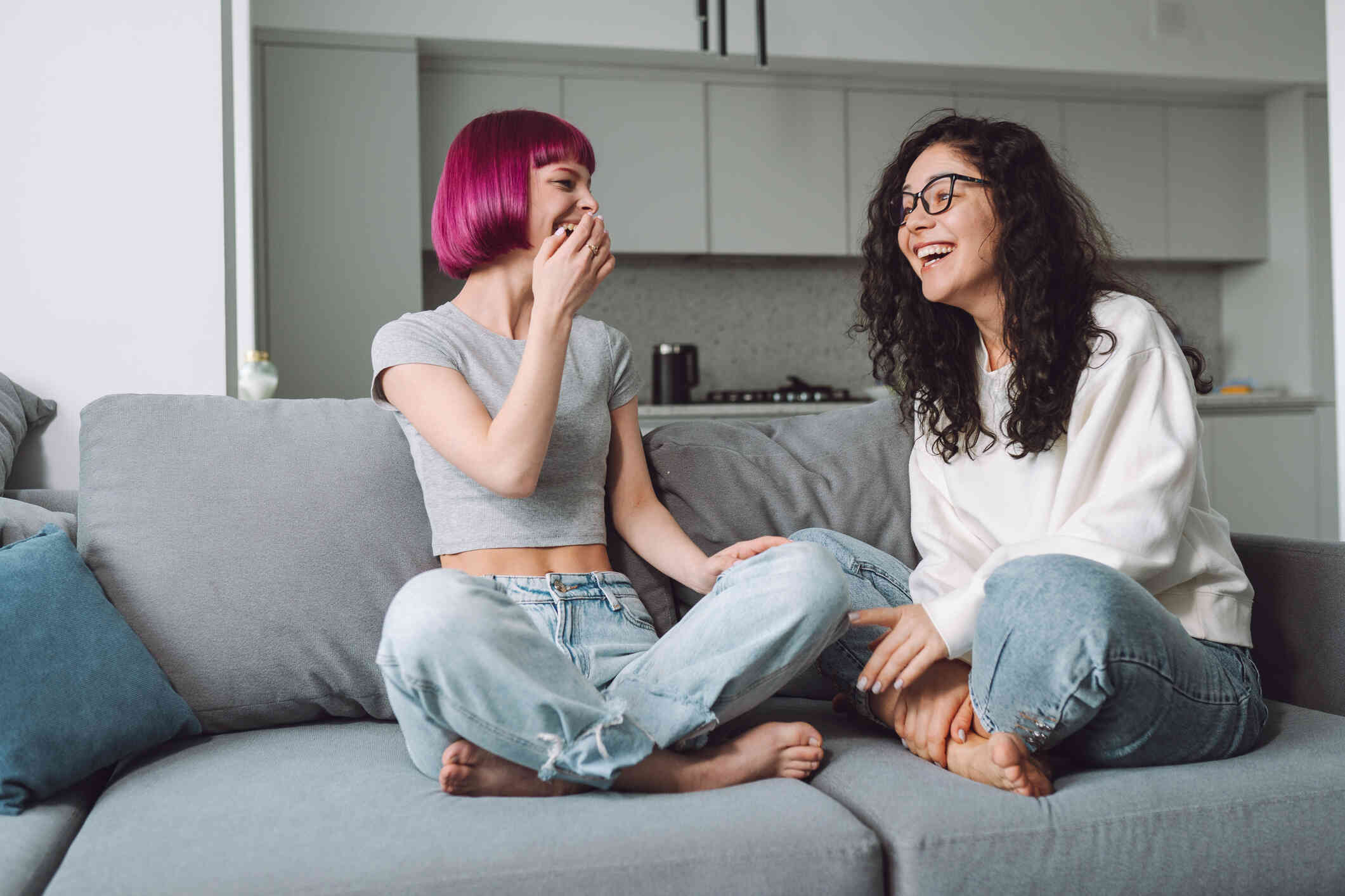 Two female friends sit cross legged on the livignr oom couch while laughing.