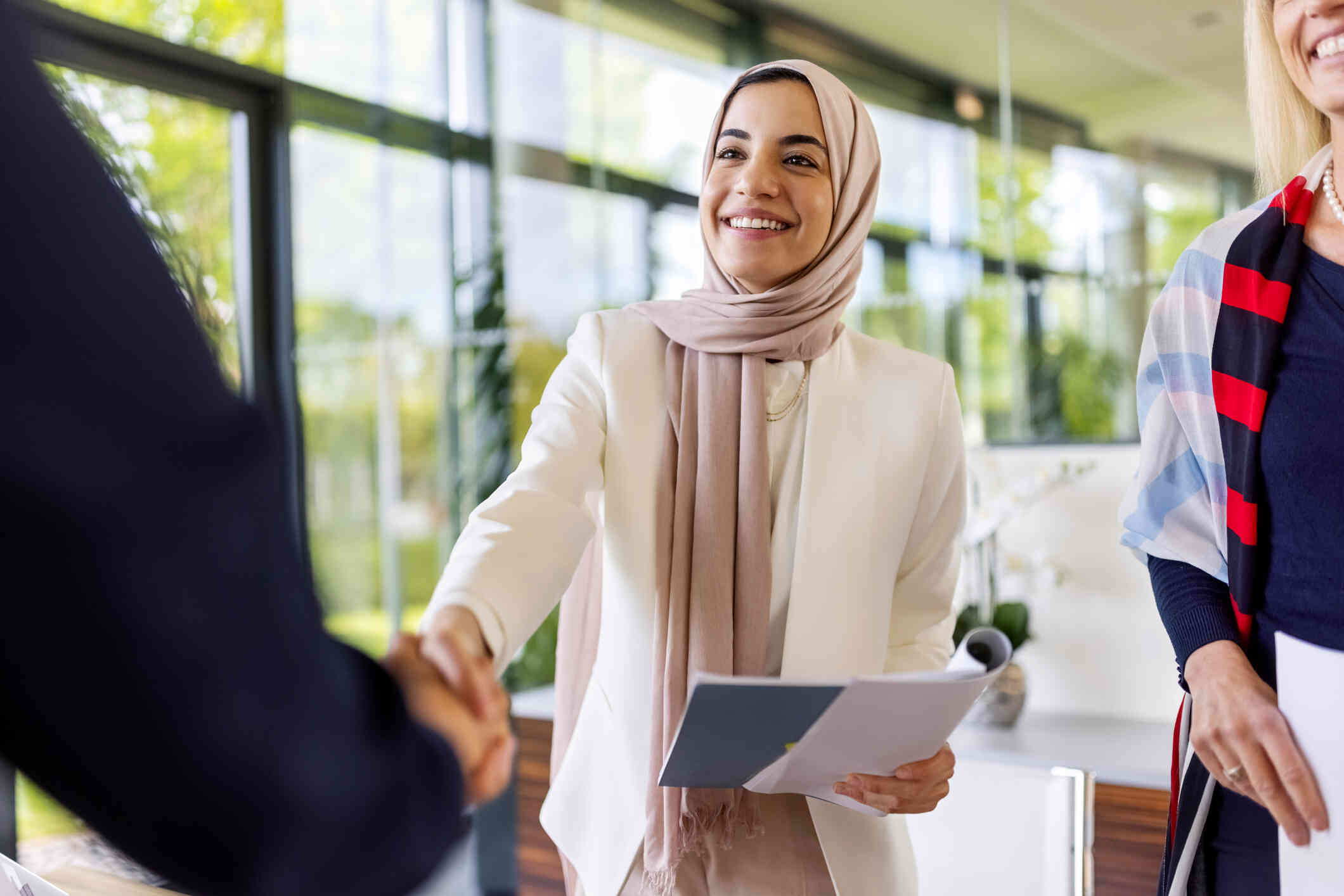 A woman wearing a pink hijab and white sweater smiles as she holds papers in one hand and shakes a person's hand with the other.