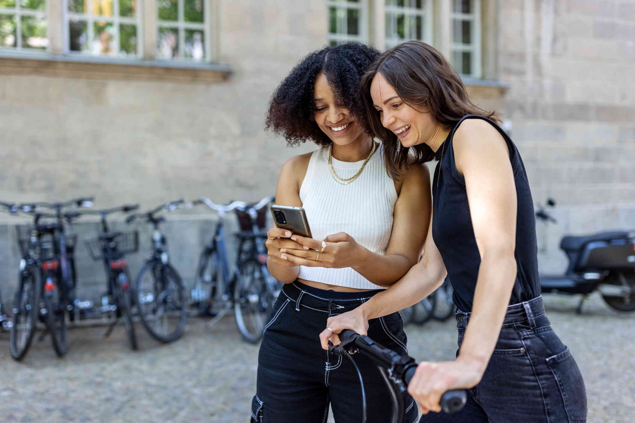 A woman with a bike stands outside next to her female friend as they smile and look at a phone screen together.