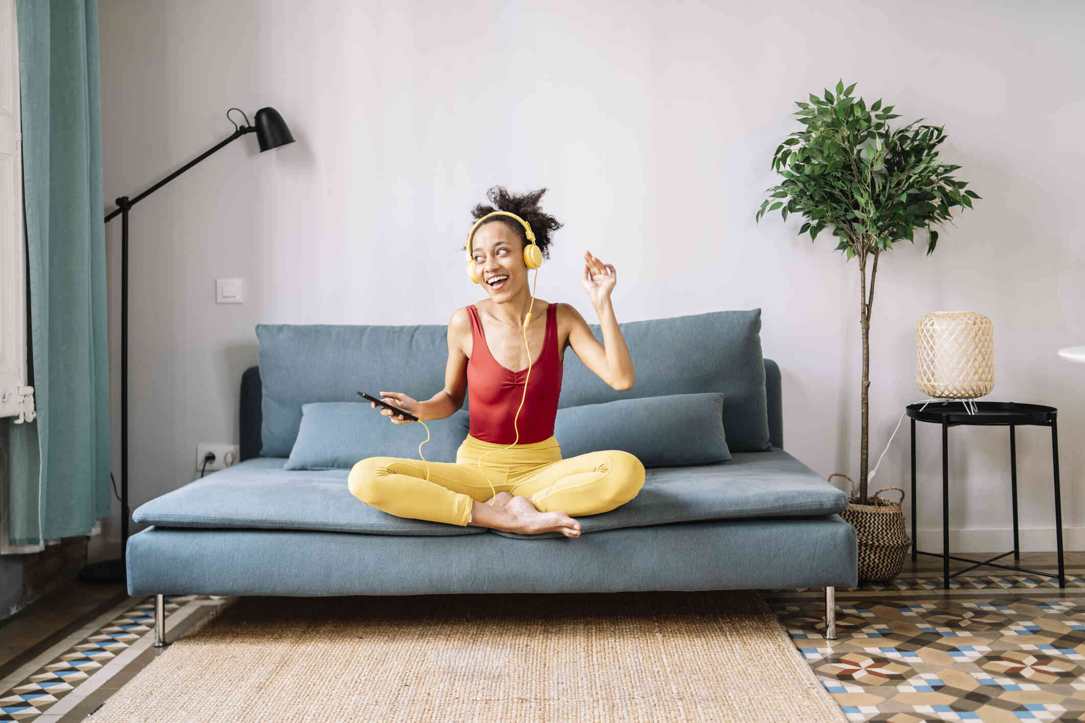 A woman ina red top and yellow pants sits cross legged on her couch and smiles brightly while listening to music with yellow headphones.