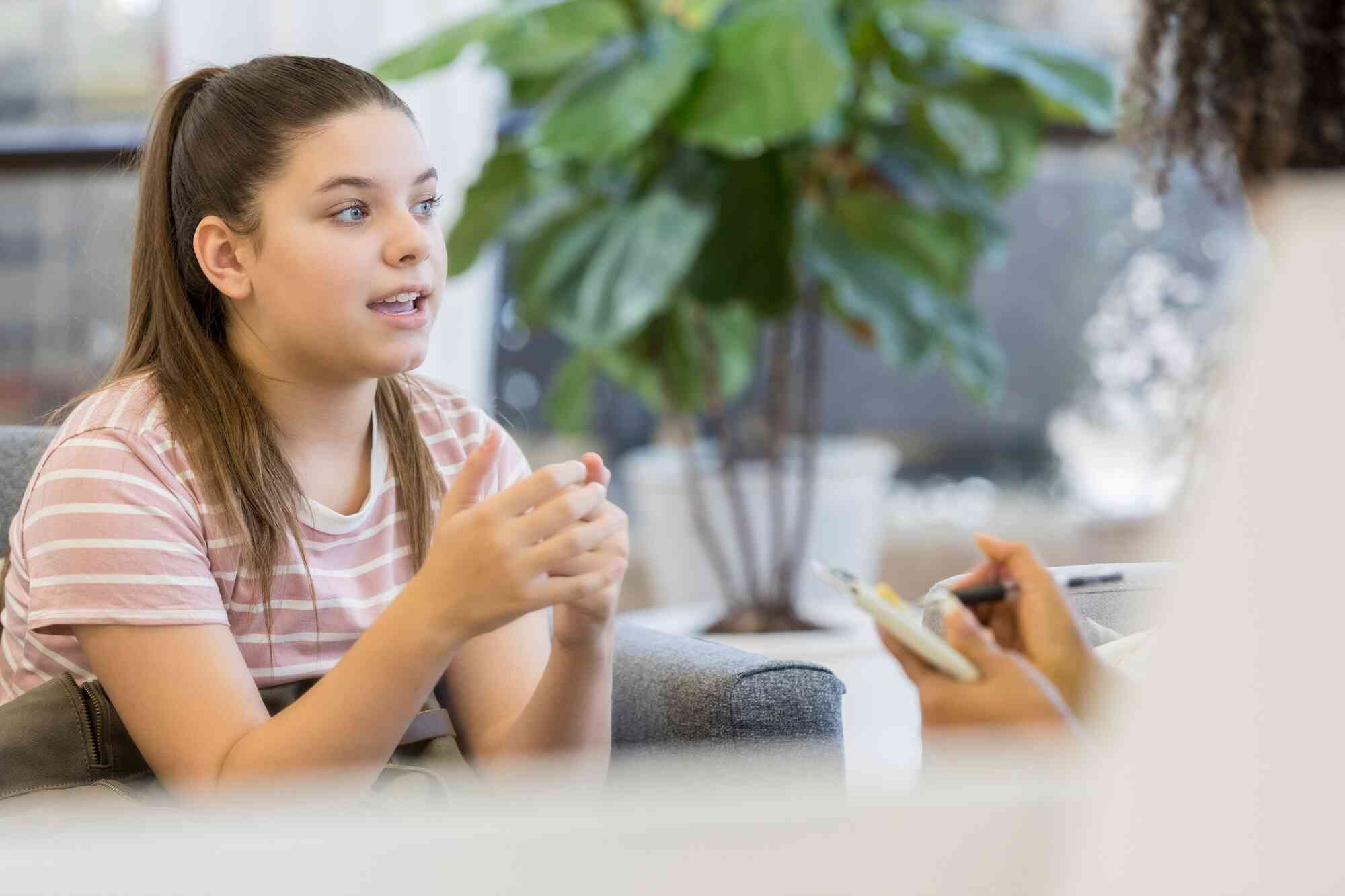 A girl in a pink striped shirt sits in a chair with a neutral expression while talking with a woman holding a clipboard