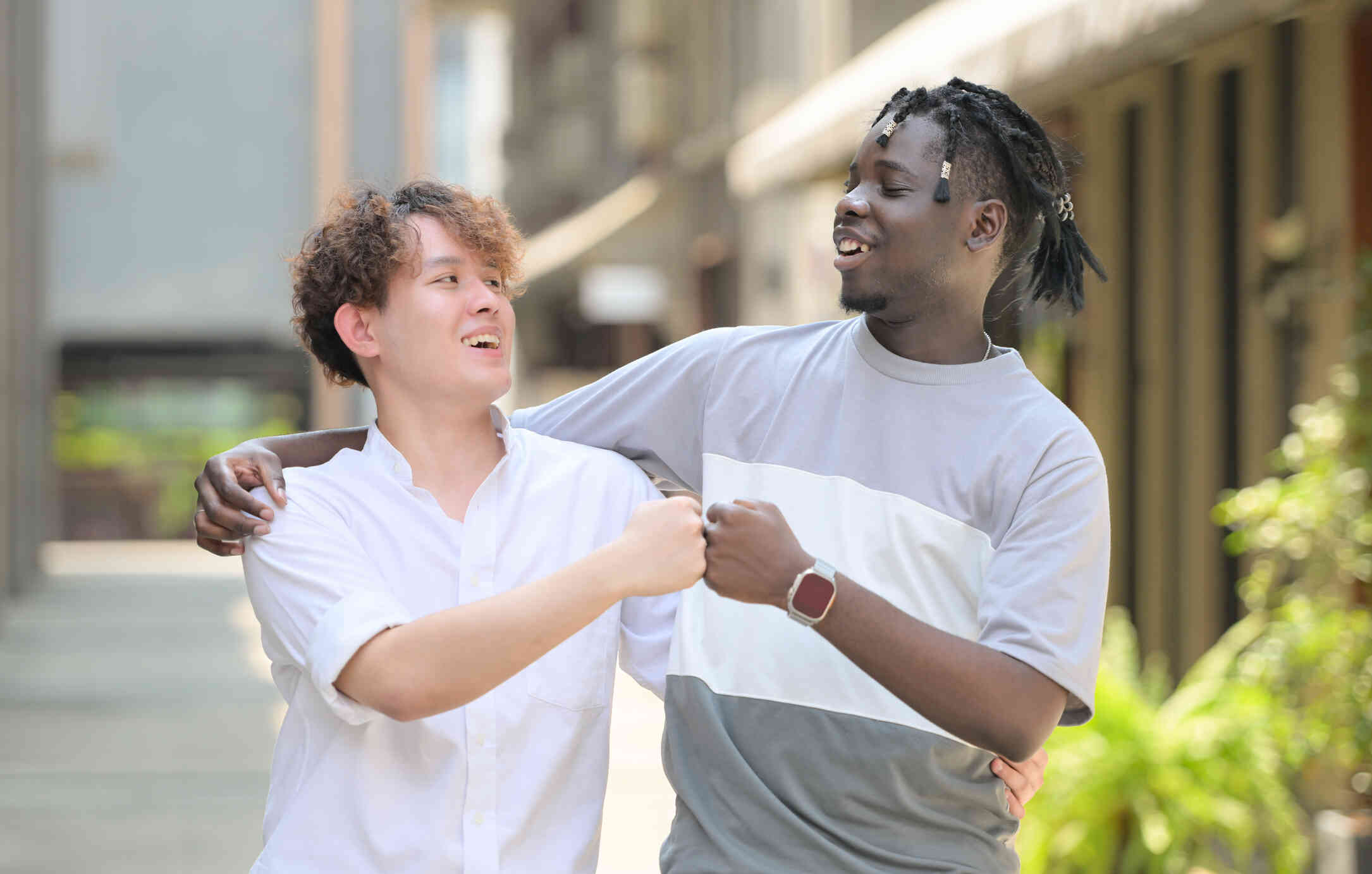 A young man in a white shirt with curly hair and a young many in a gray striped shirt with braids fist bump each other at they stand outside with an around each others shoulder.