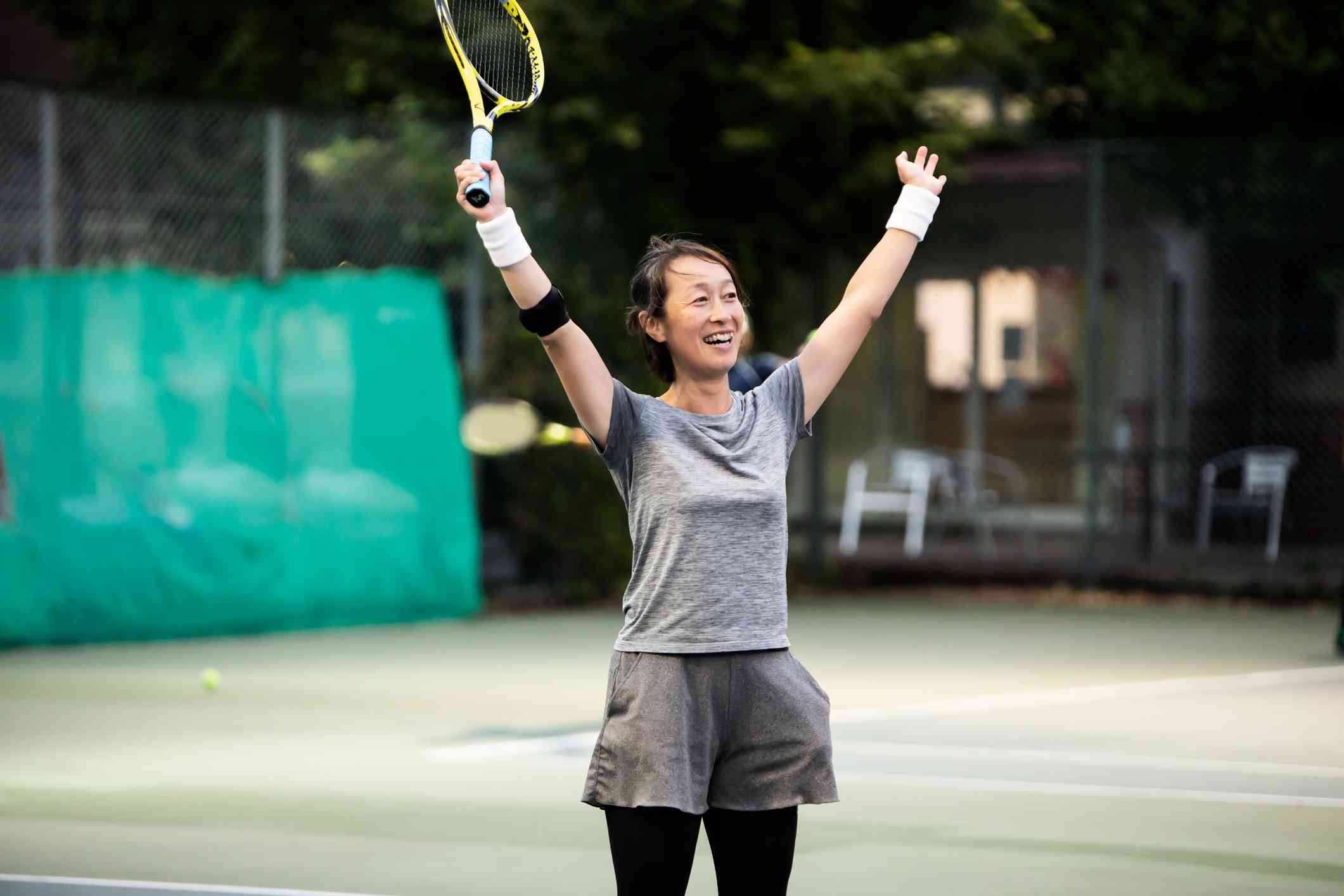 A woman in a grey shirt smiles and raises her arms in victory while holding a tennis racket and standing on a tennis court.