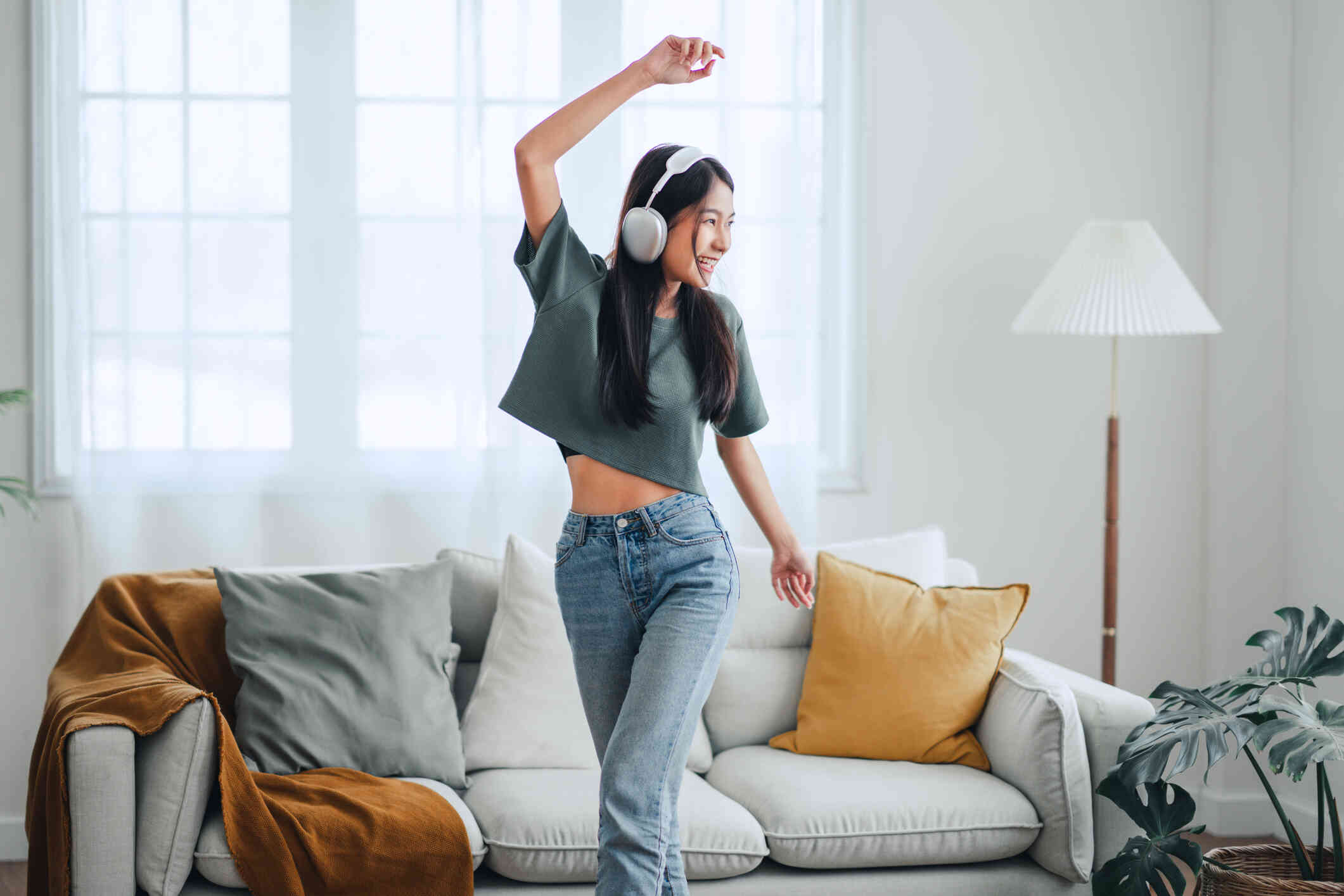 A young woman wearing jeans, a t-shirt, and headphones smiles as she dances around in a living room.