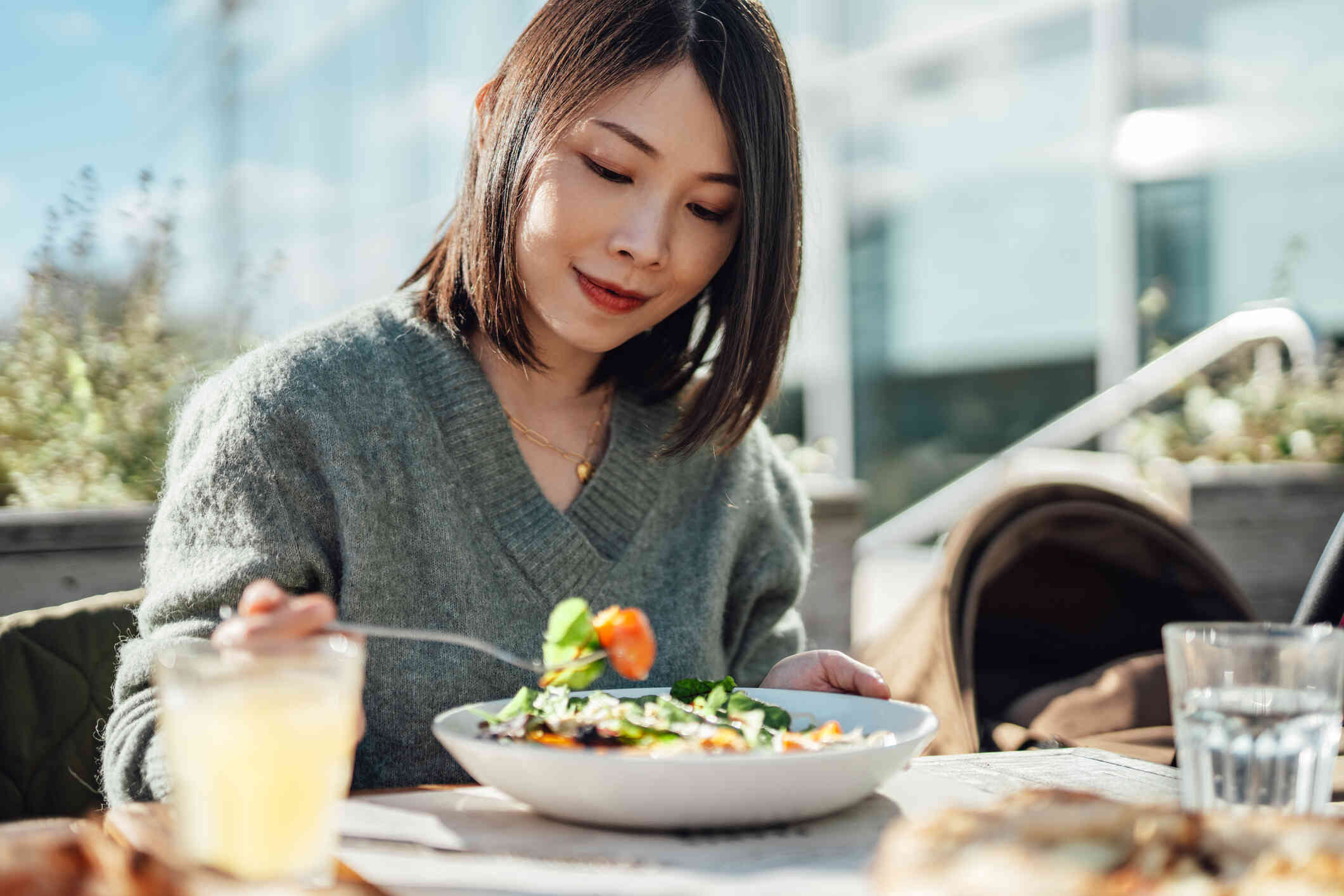 A woman in a gray sweater sits outside at a table and picks up a bite of food from a bowl in front of her using a fork.