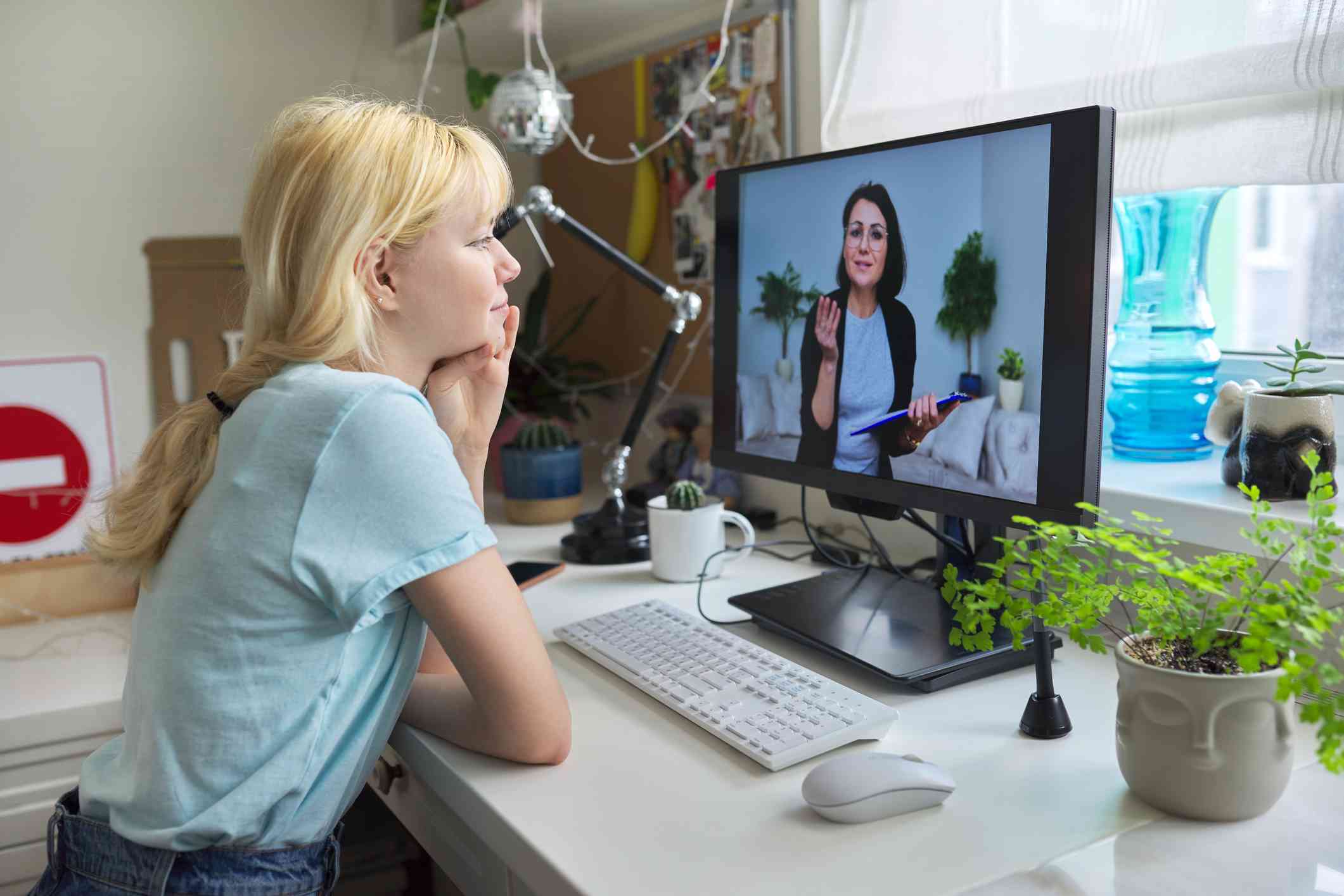 A teenage girl with blonde hair and a blue shirt sits at her desk and looks at her computer where a therapist is speaking with her online.