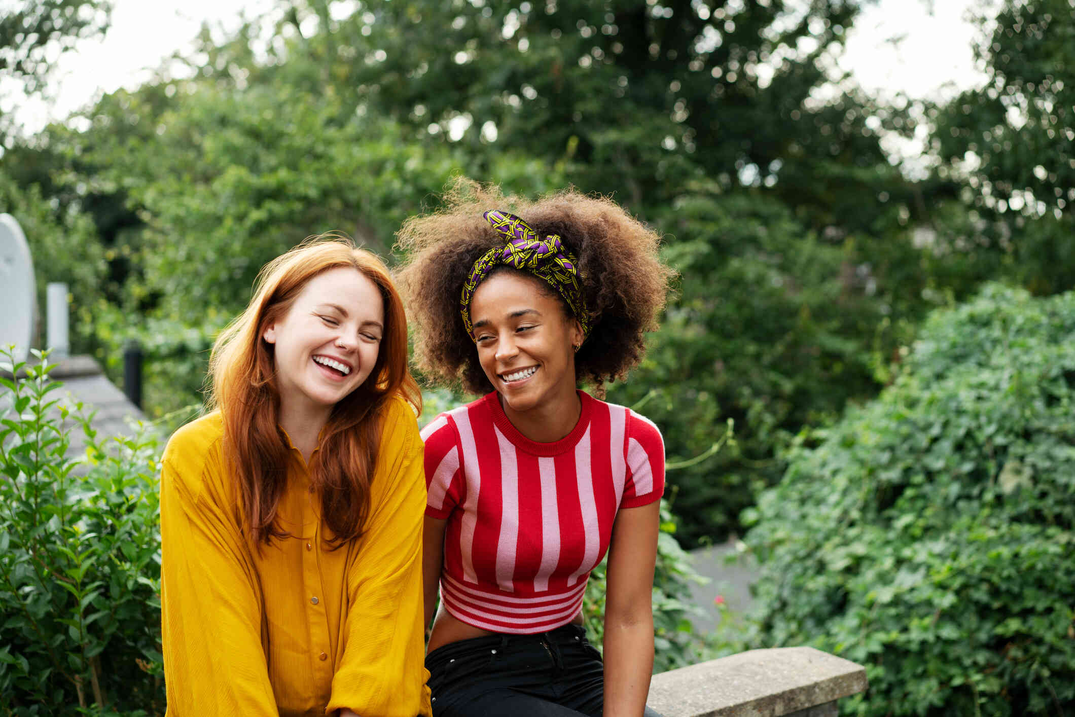 Two adult female friends sit next to each other outside on a sunny day while smiling.