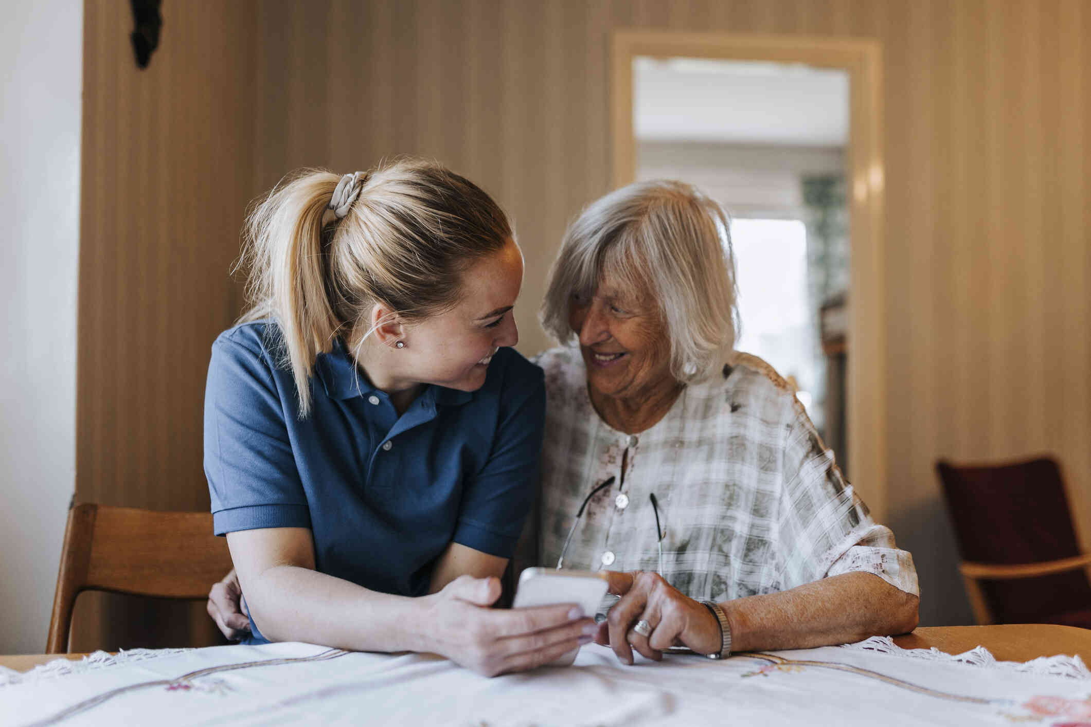 An elderly woman and a young woman relative sit close together at the kitchen table as the young woman holds a phone in her hand they the two woman smile at each other.