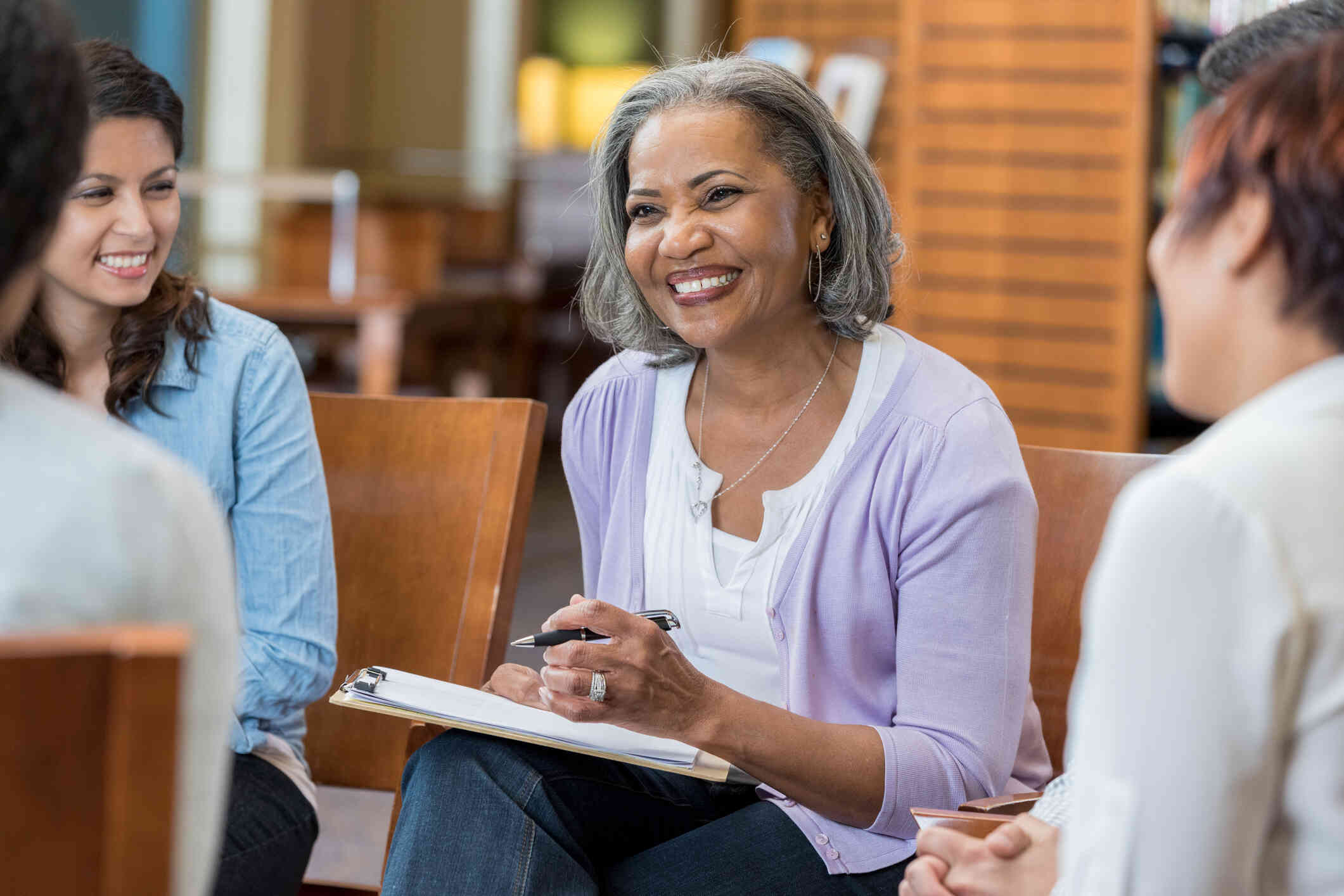 A middle aged female therapist in a purple cardigan smiles while holding a clipboard and sitting in a group therapy circle.