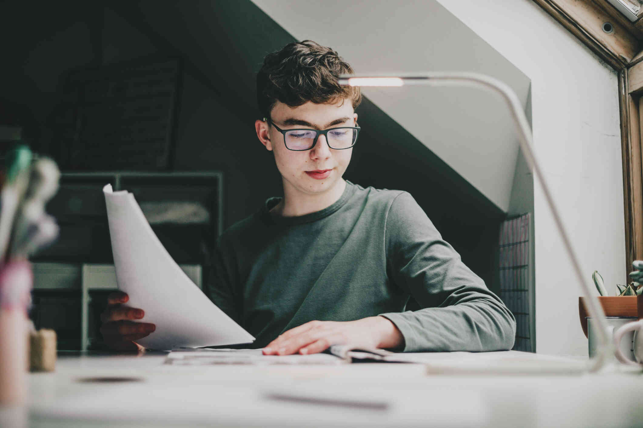 A teenage boy ists at his desk in a darker lit room while holding papers and looking at his computer