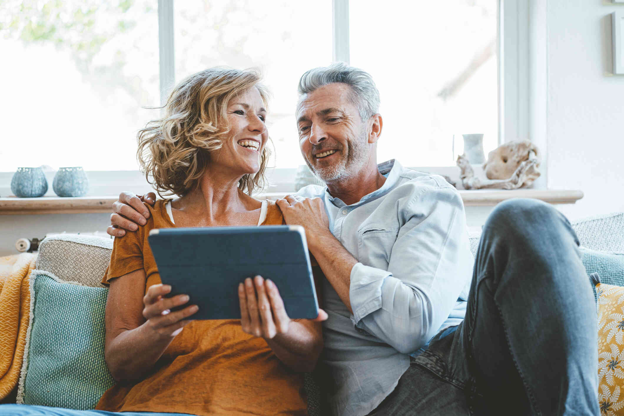 A woman with short blonde hair smiles as she holds a tablet and sits next to a man with gray hair who is smiling and holding her shoulders.