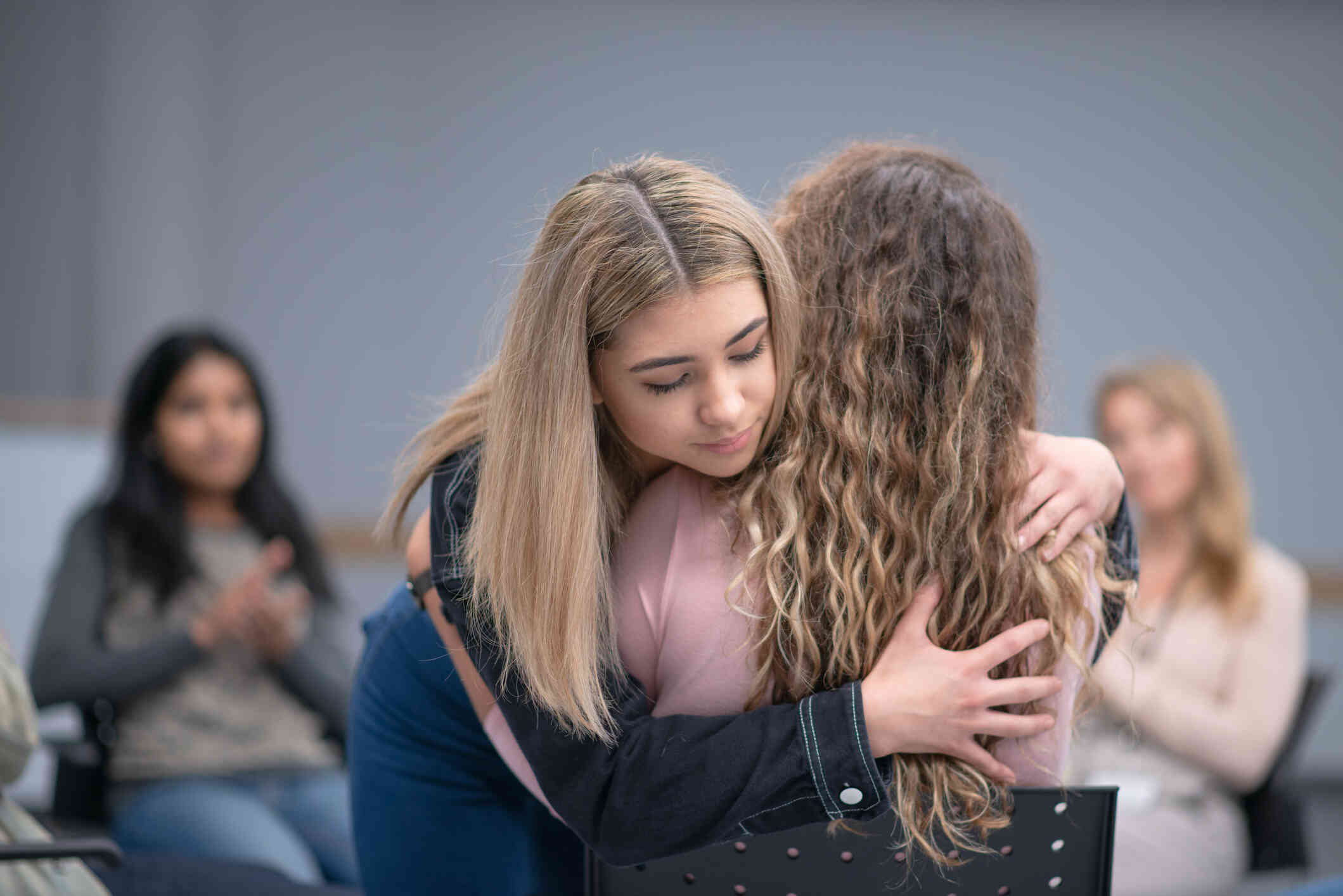 Two young girls hugging, with other teenagers visible blurred in the background.