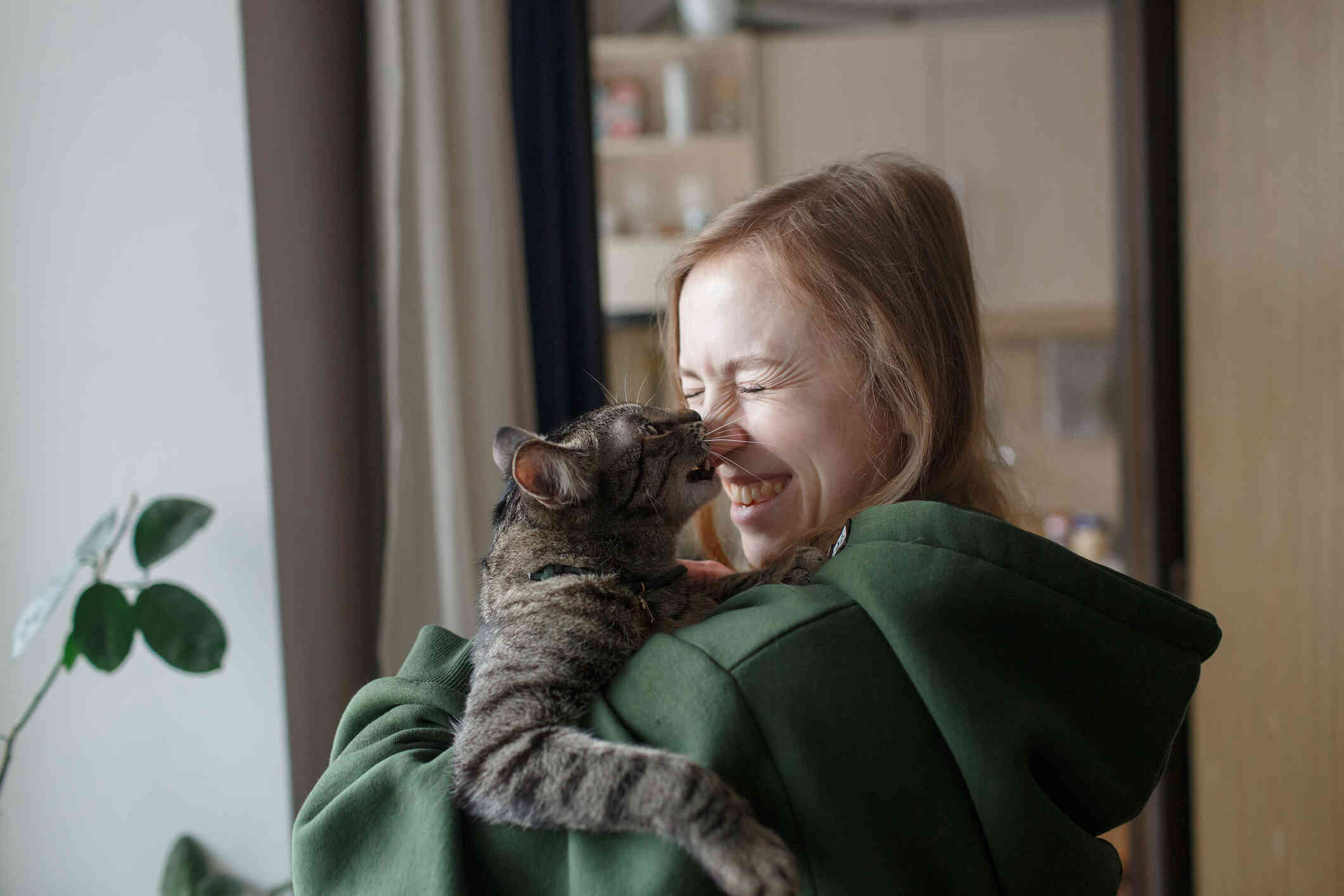 A close up of a woman in a green shirt holding her cat close to her face while smiling and the cat nibbles her nose.