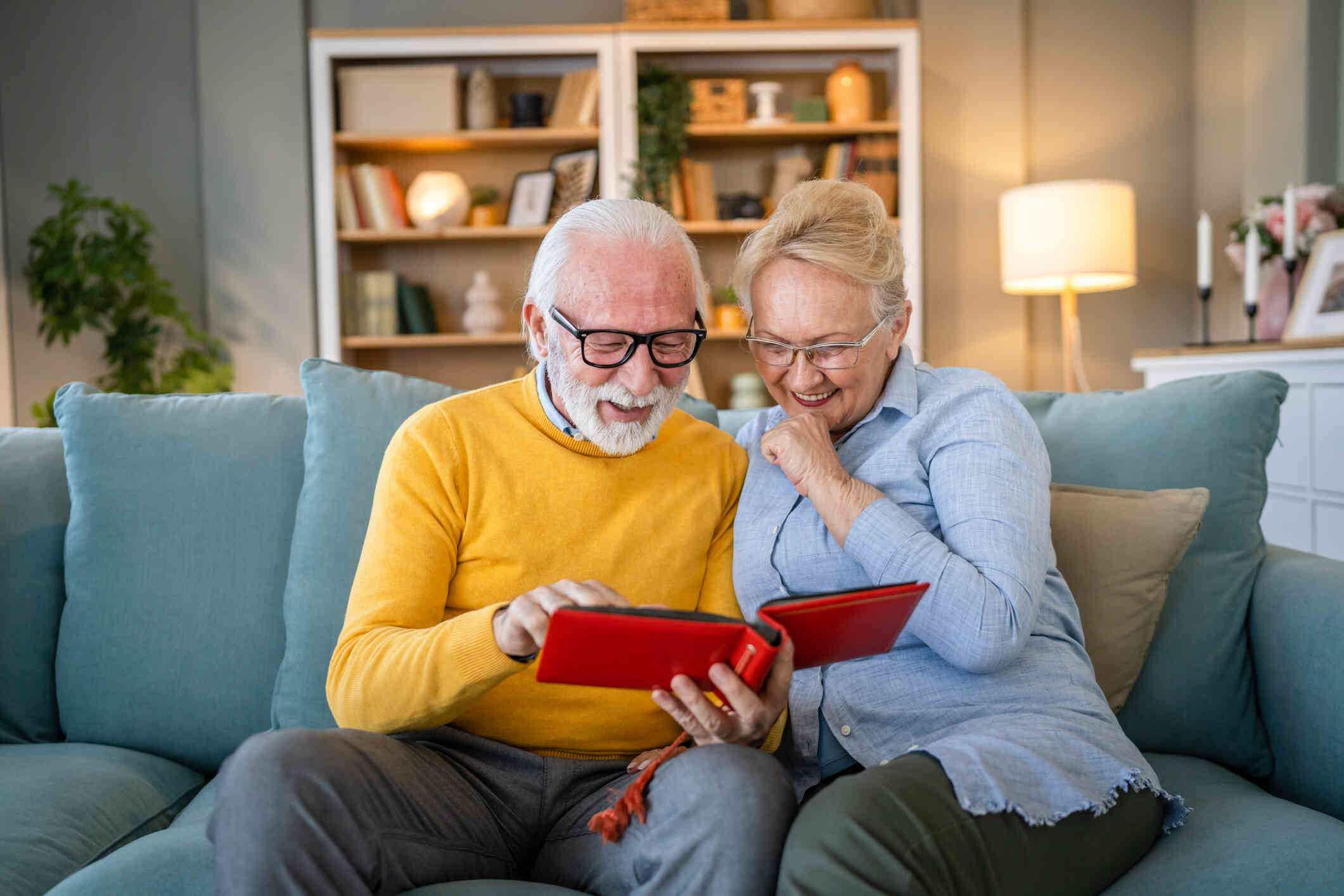 An elderly male and female couple sit side by side on the couch and look at a red photo album together while smiling.