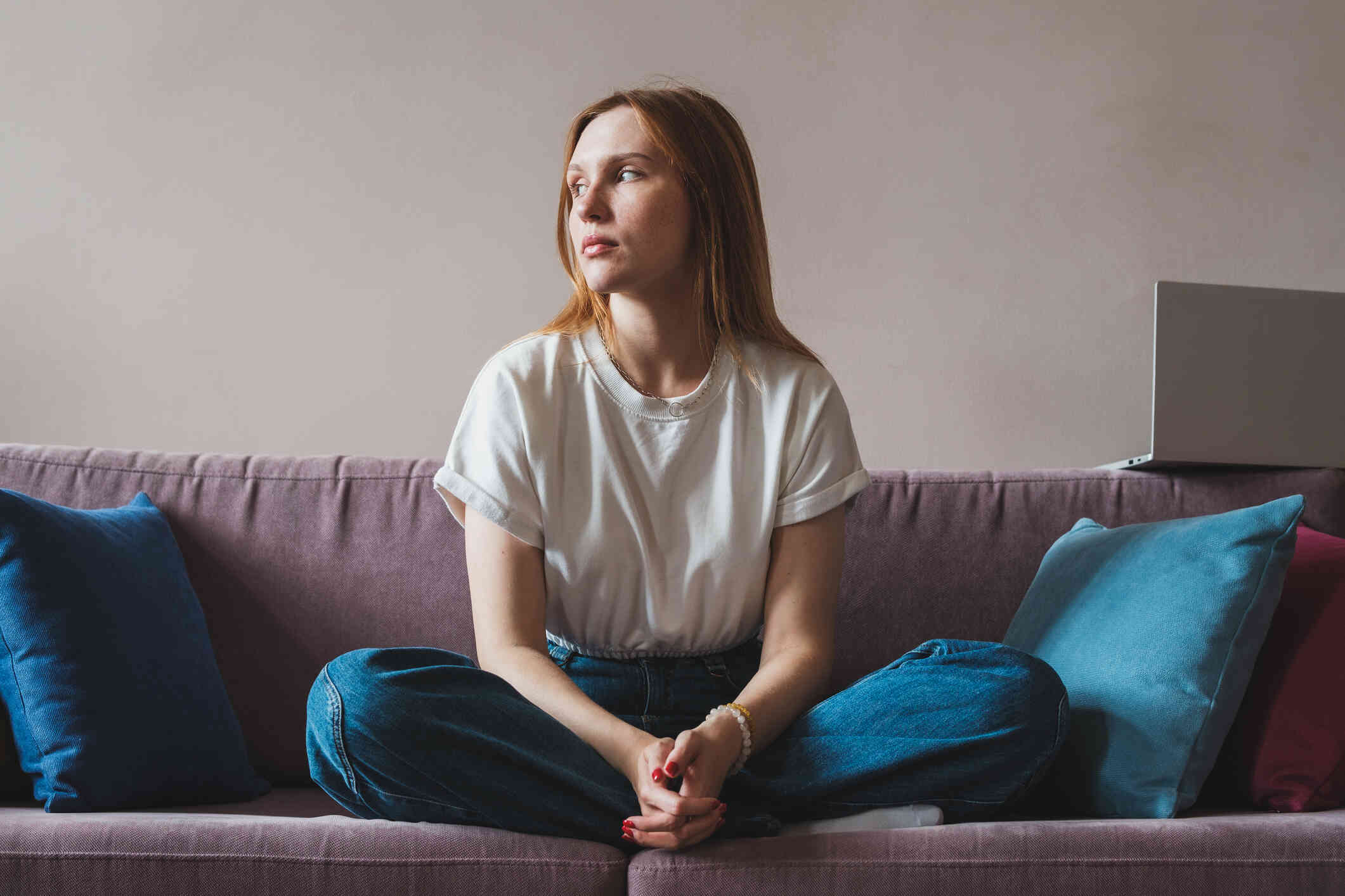 A woman in a white shirt sits cross legged on the couch and gazes off with a sad expression.