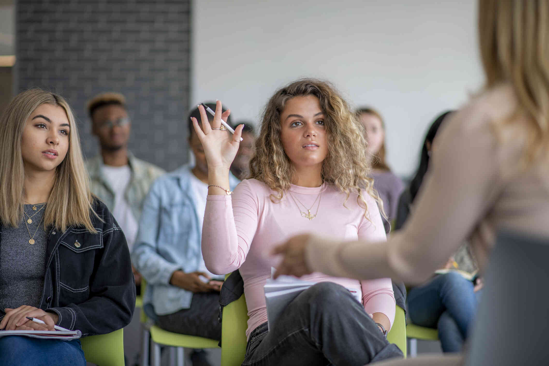 A woman with curly, blonde hair wears a pink sweater and raises her hand as she sits in a class. Another woman stands in front of the the class speaking.