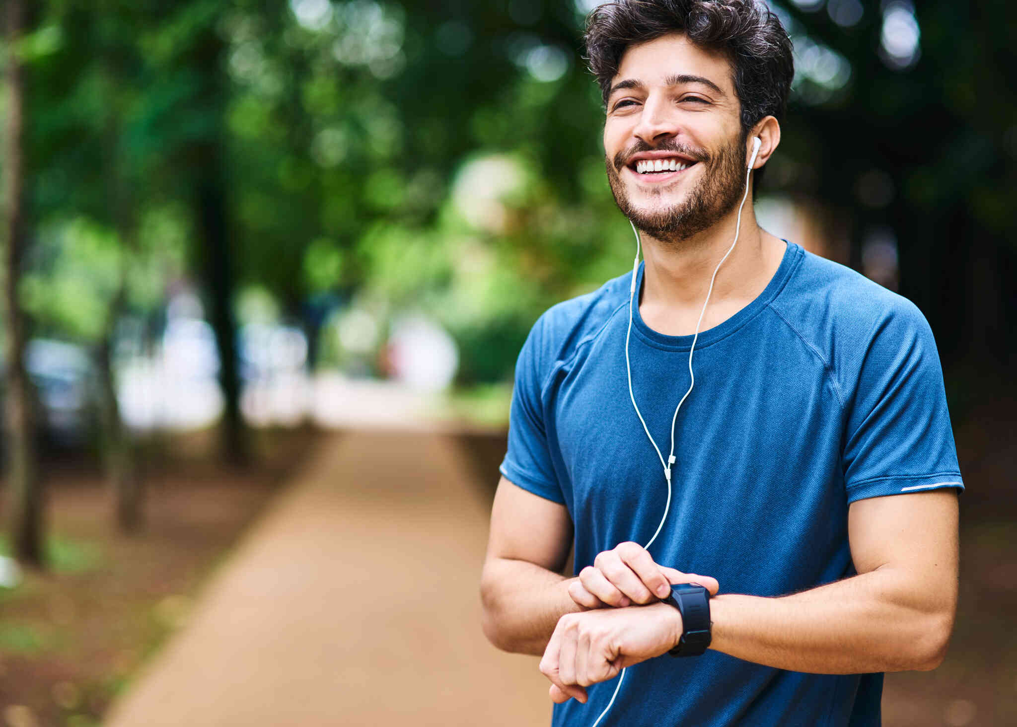 A man in a blue shirt with headphones stands outside on a sunny day and smiles brightly while tapping on his smart watch.