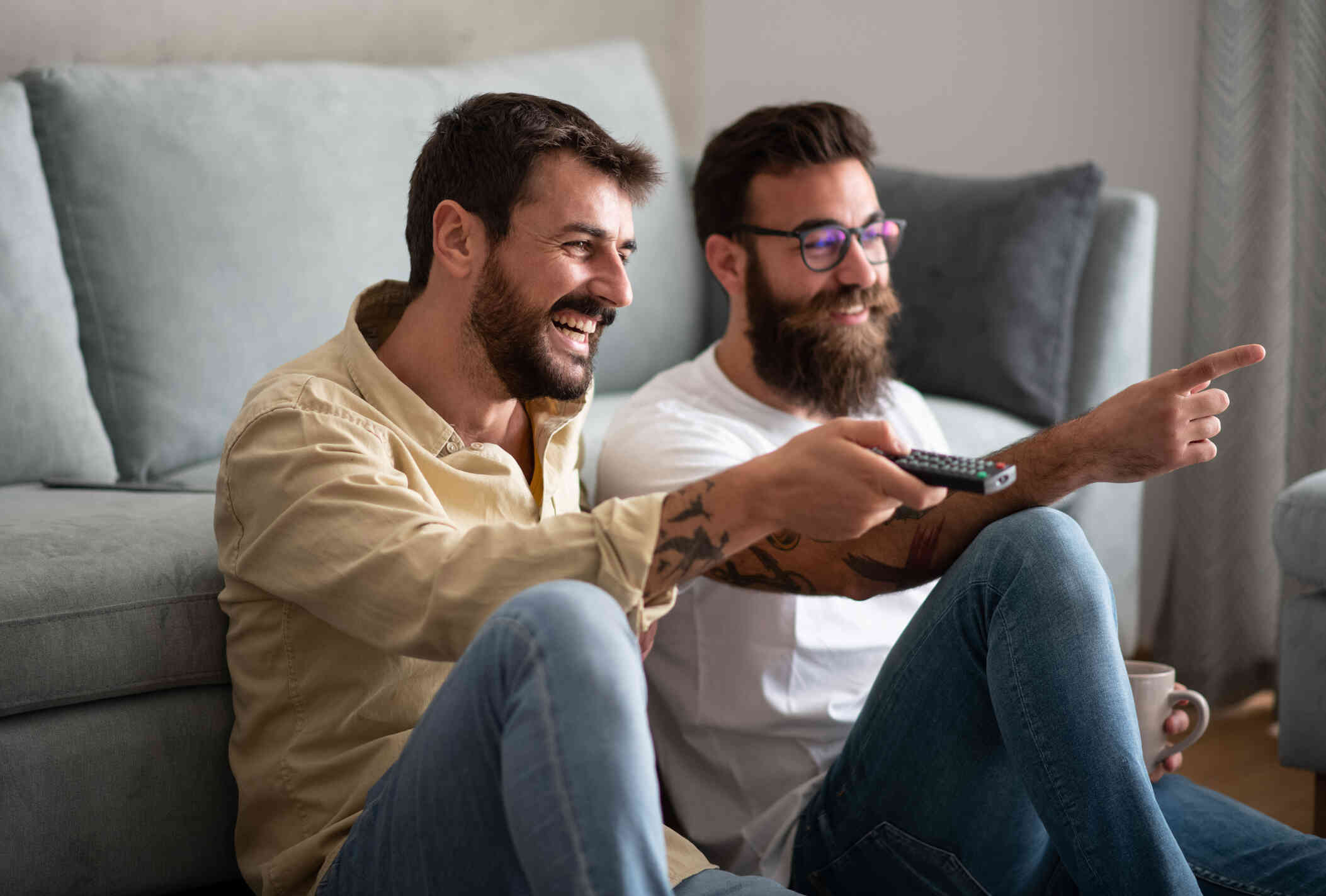 Two male friends sit happily on the living room floor as one holds a tv remote in his hand.