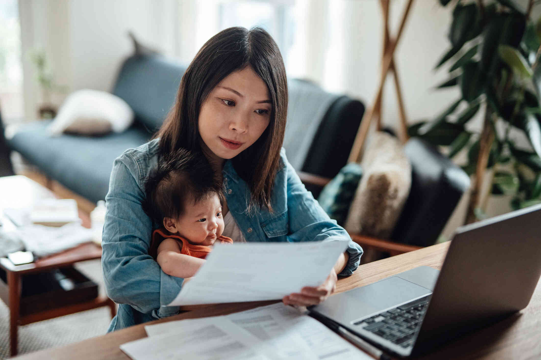 A woman sits at  table with her newborn baby and looks at a piece of paper with a serious expression.