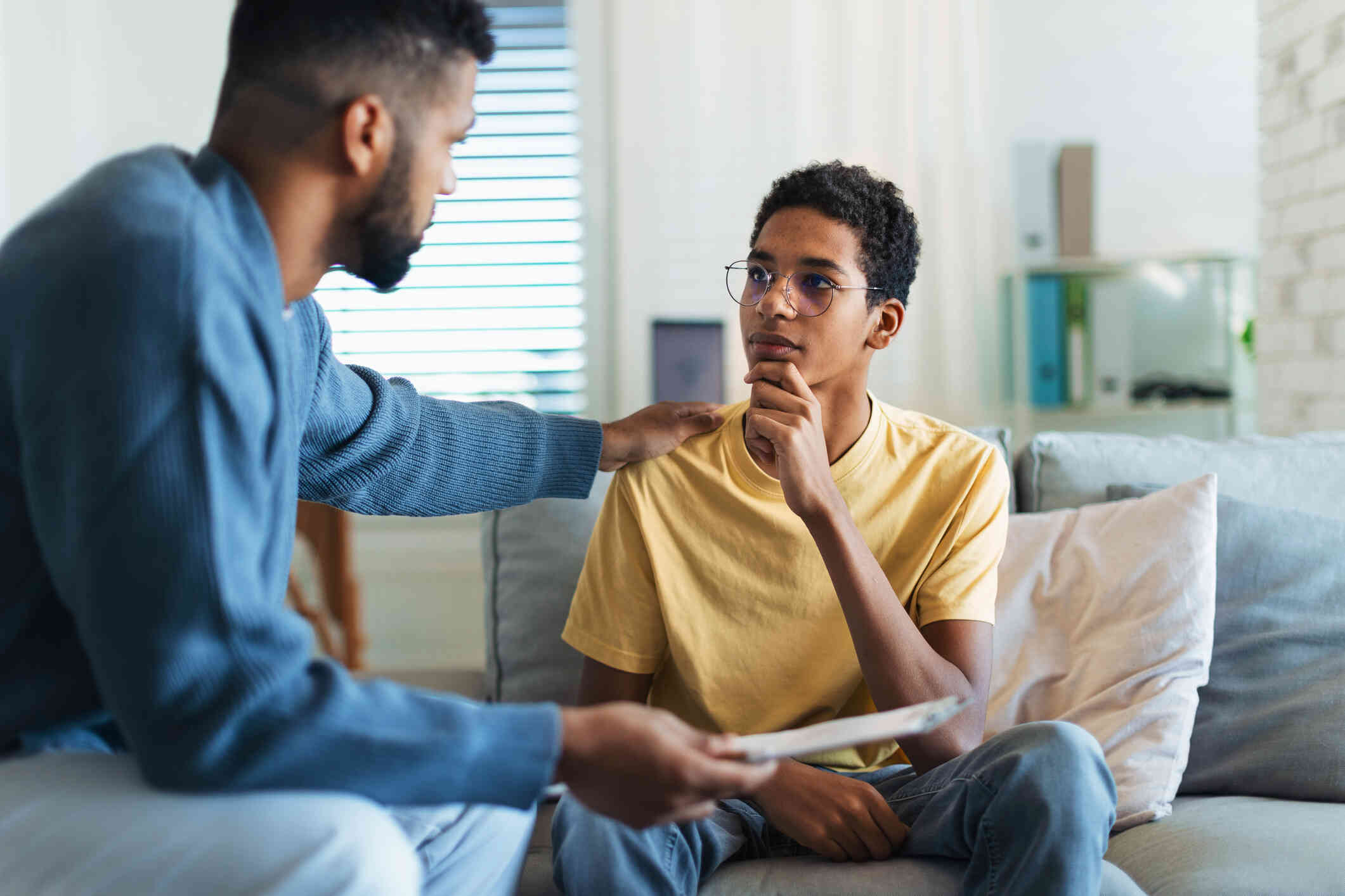 A male therapist in a blue shirt rests his hand on a his young male pateint shoulder during a therapy sesson.