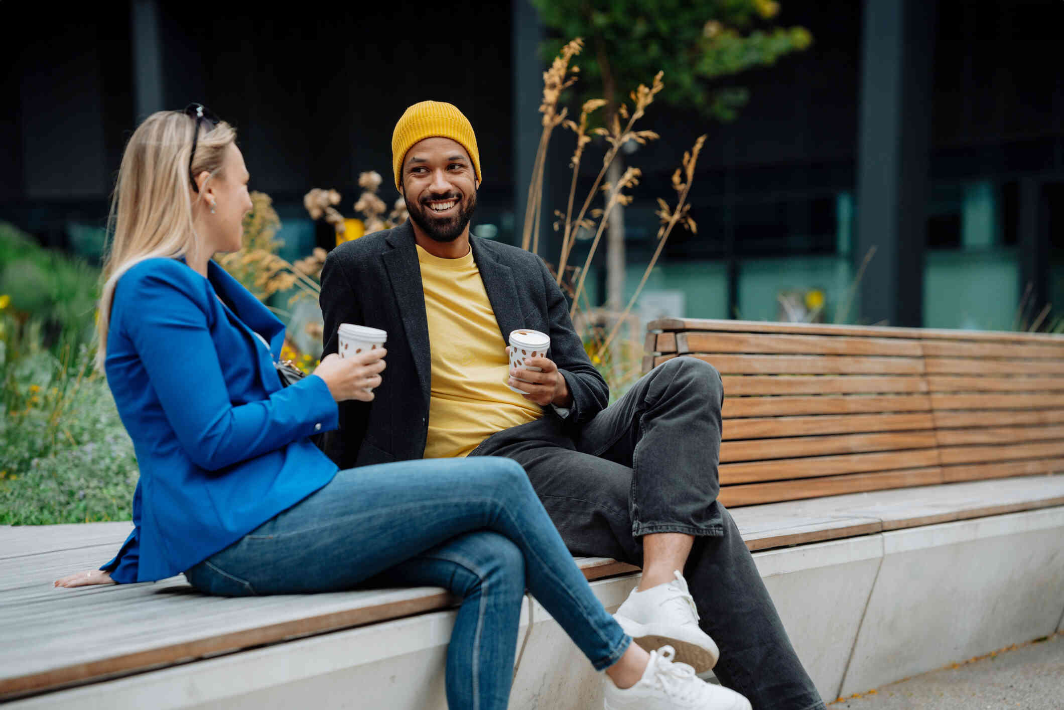 A woman and her male friend sit next to each other on a park bench while drinking cup of coffee and chatting.