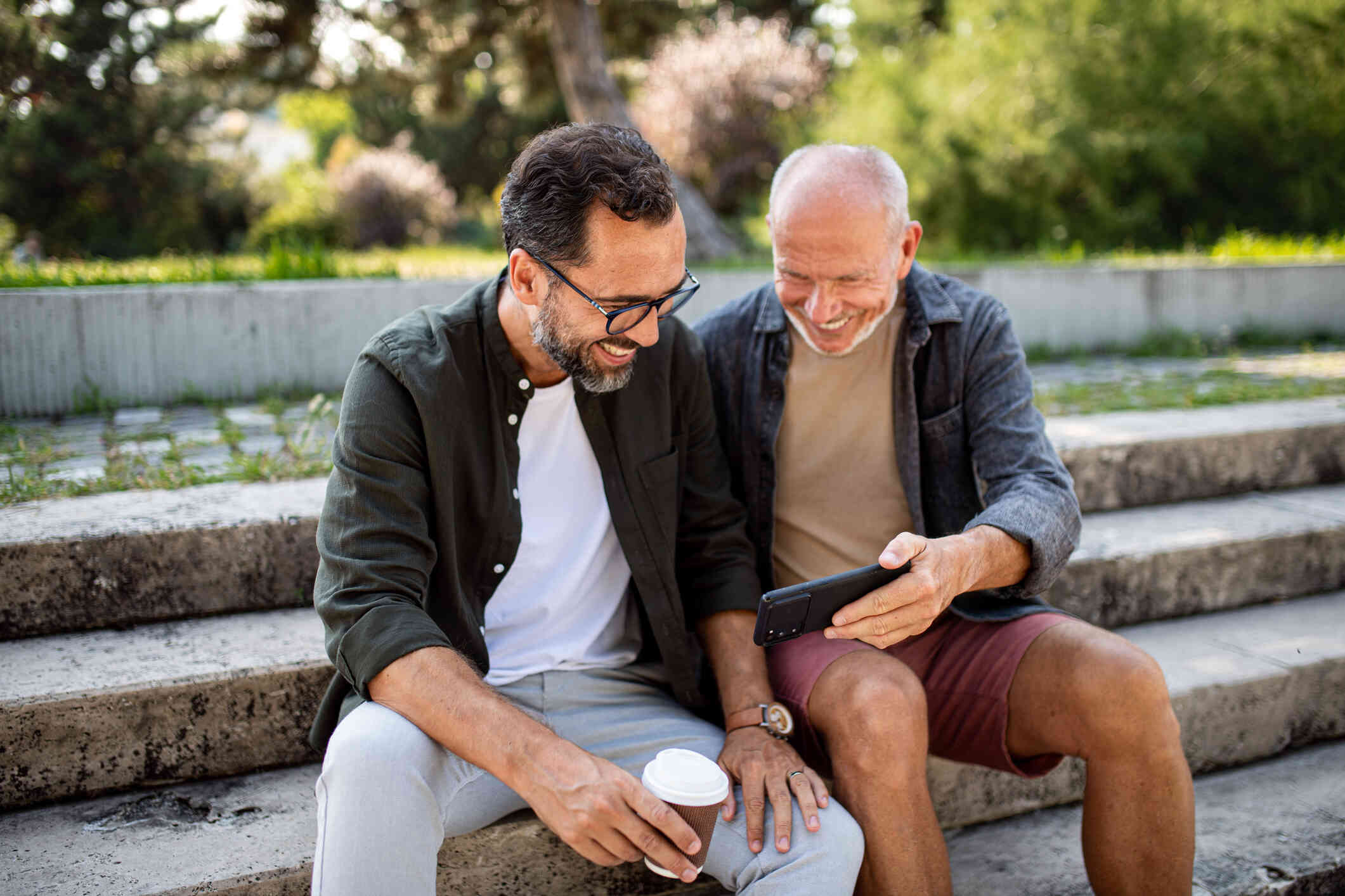 Two middle aged male friends sit on a cement step outside on a sunny day while looking at a phone screen together.