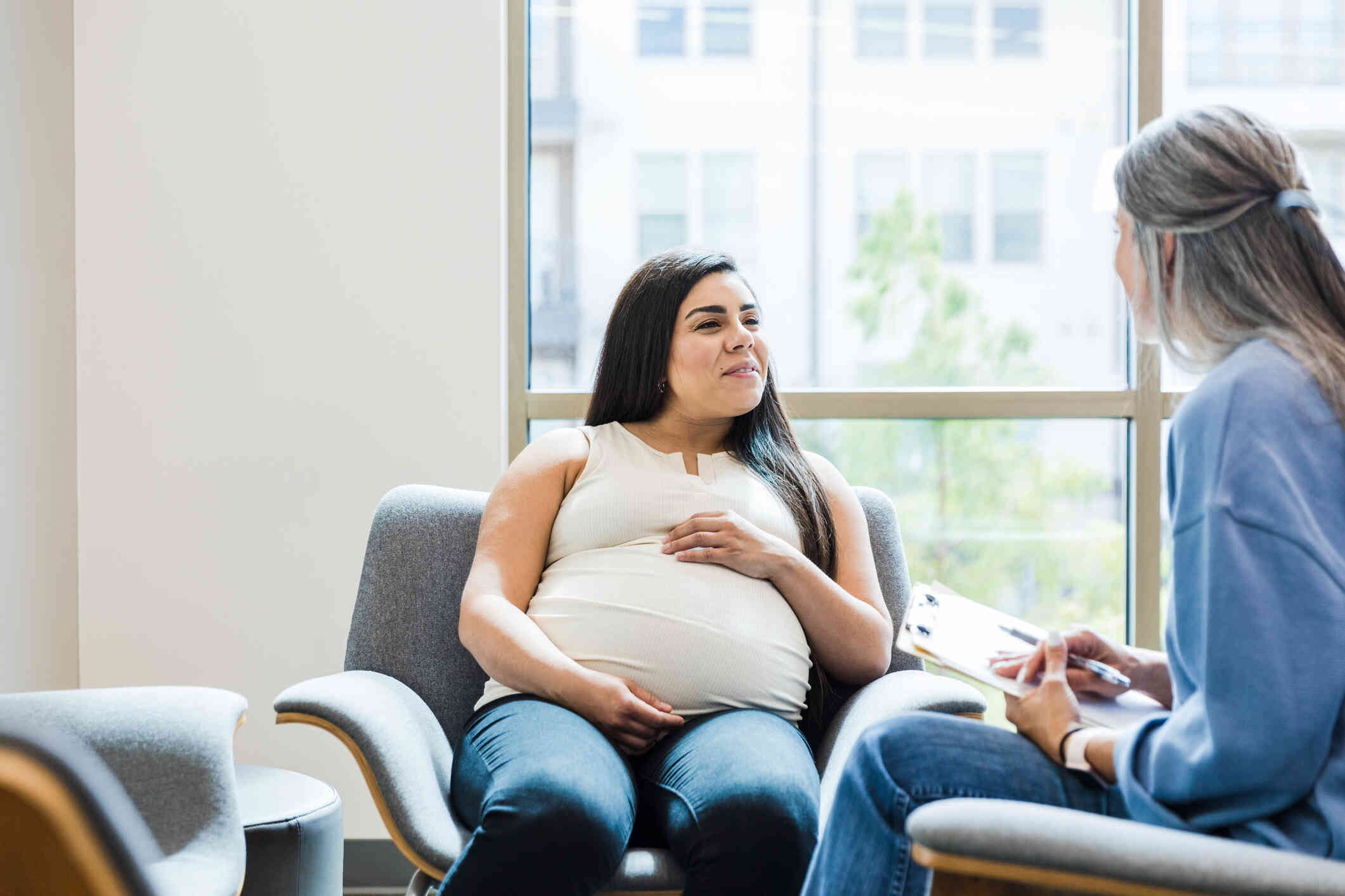 A pregnant woman sits in an armchair  and smiles at the female therapist who sits across from her.