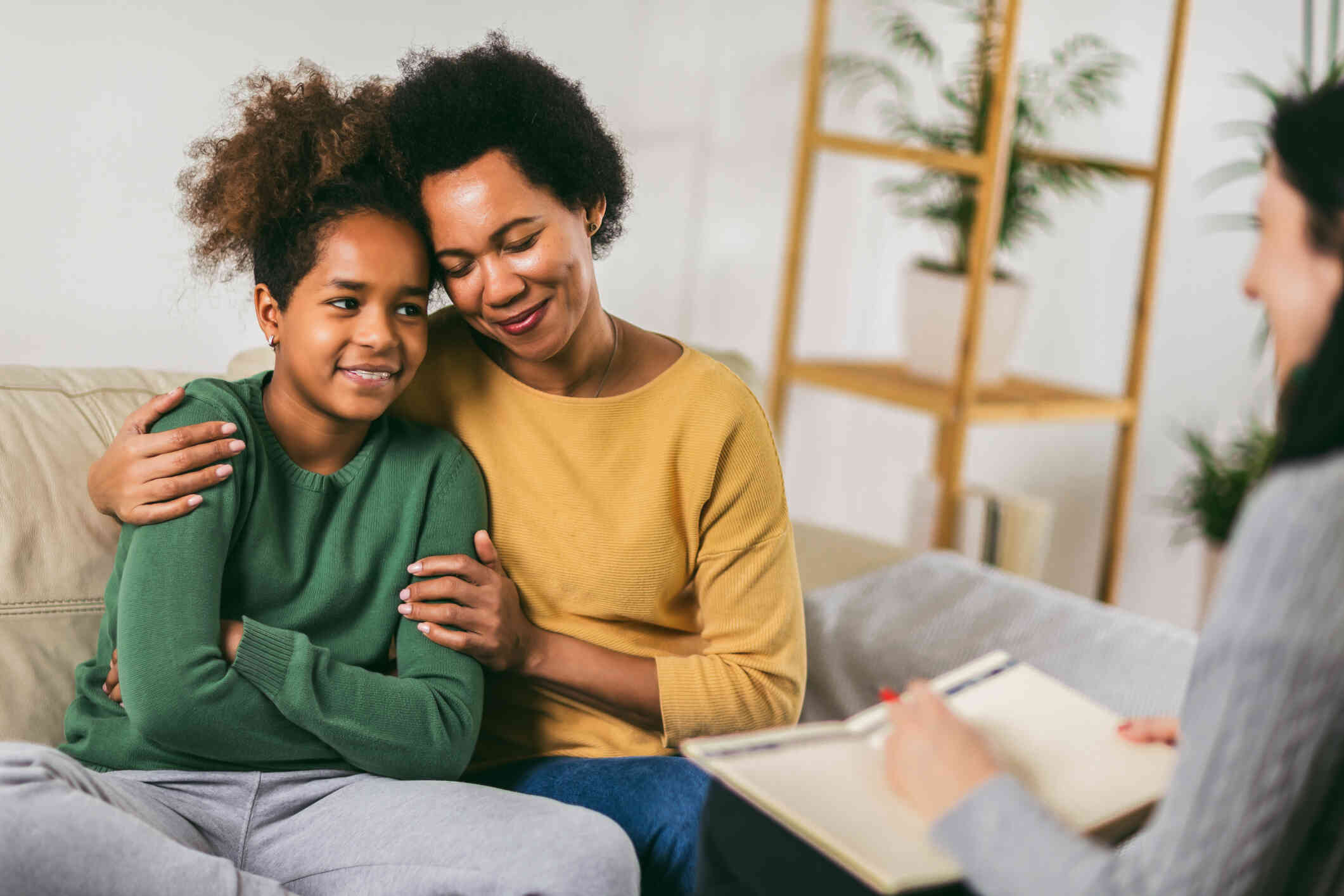 A mothers wraps her arms around her daighter as they both smile softly while sitting on a couch across from a therapist.