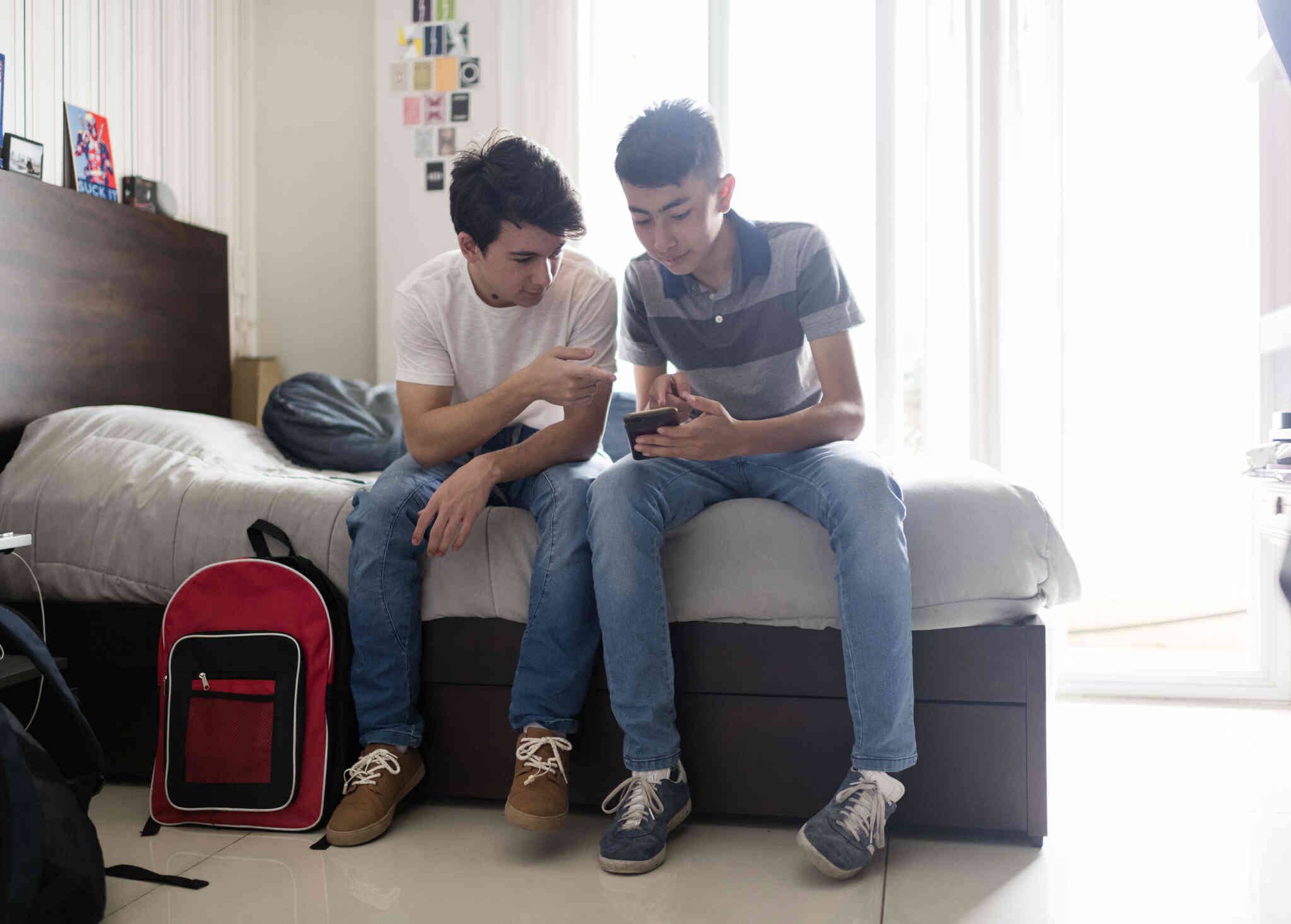 A teenage boy in a white shirt and a teenage boy in a grey striped shirt sit on a bed while looking at the phone