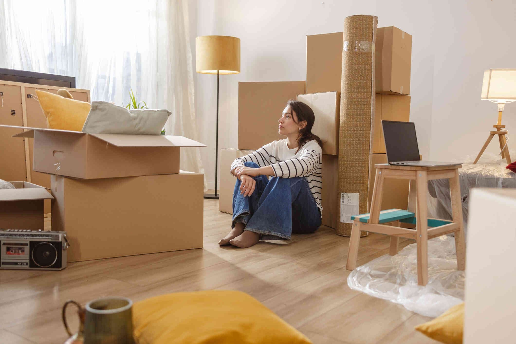 A woman n a striped sweater sits on the living room floor in a room full of moving boxes as she gazes off.