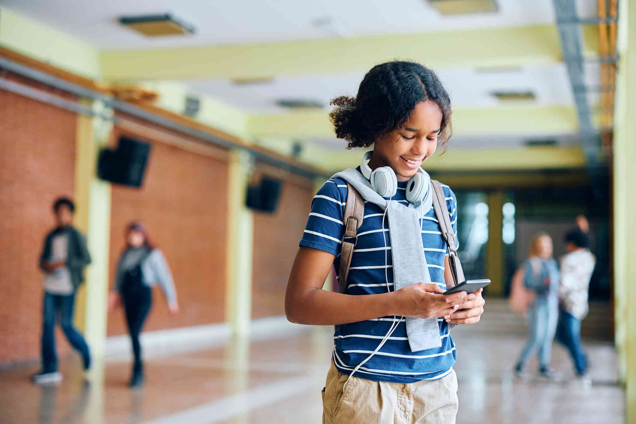 A teenage girl in a blue shirt with white headphones stands in the school hallways and looks down at the cellphone in her hand.