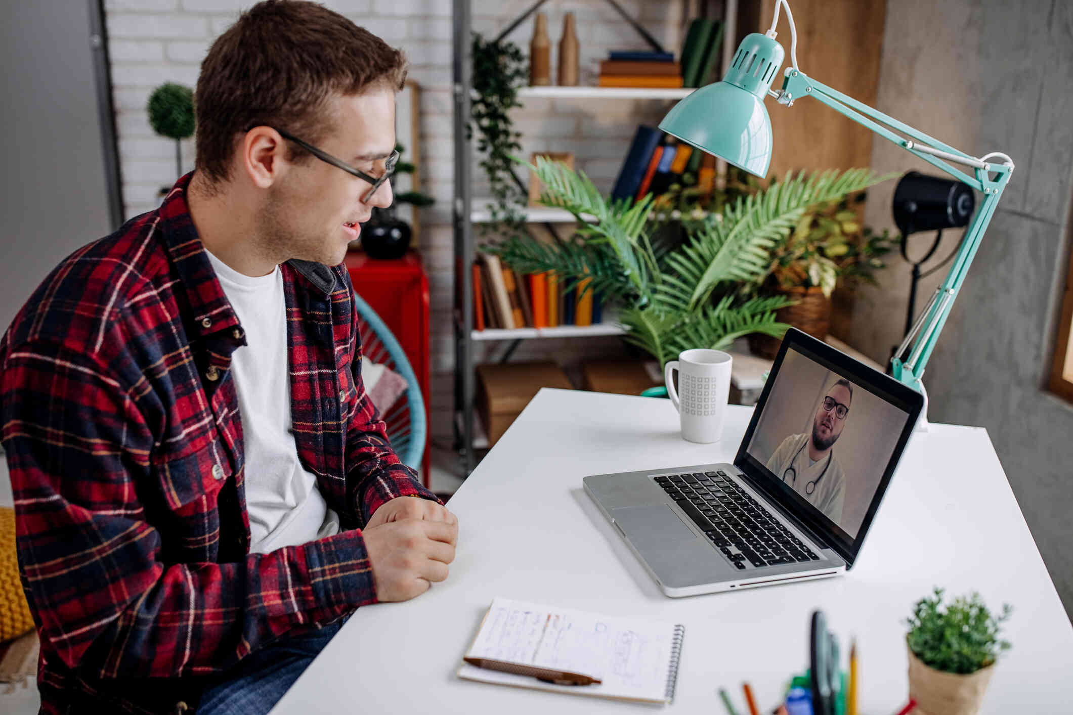 A young man in a plaid shirt having a video call on his laptop in a home office.