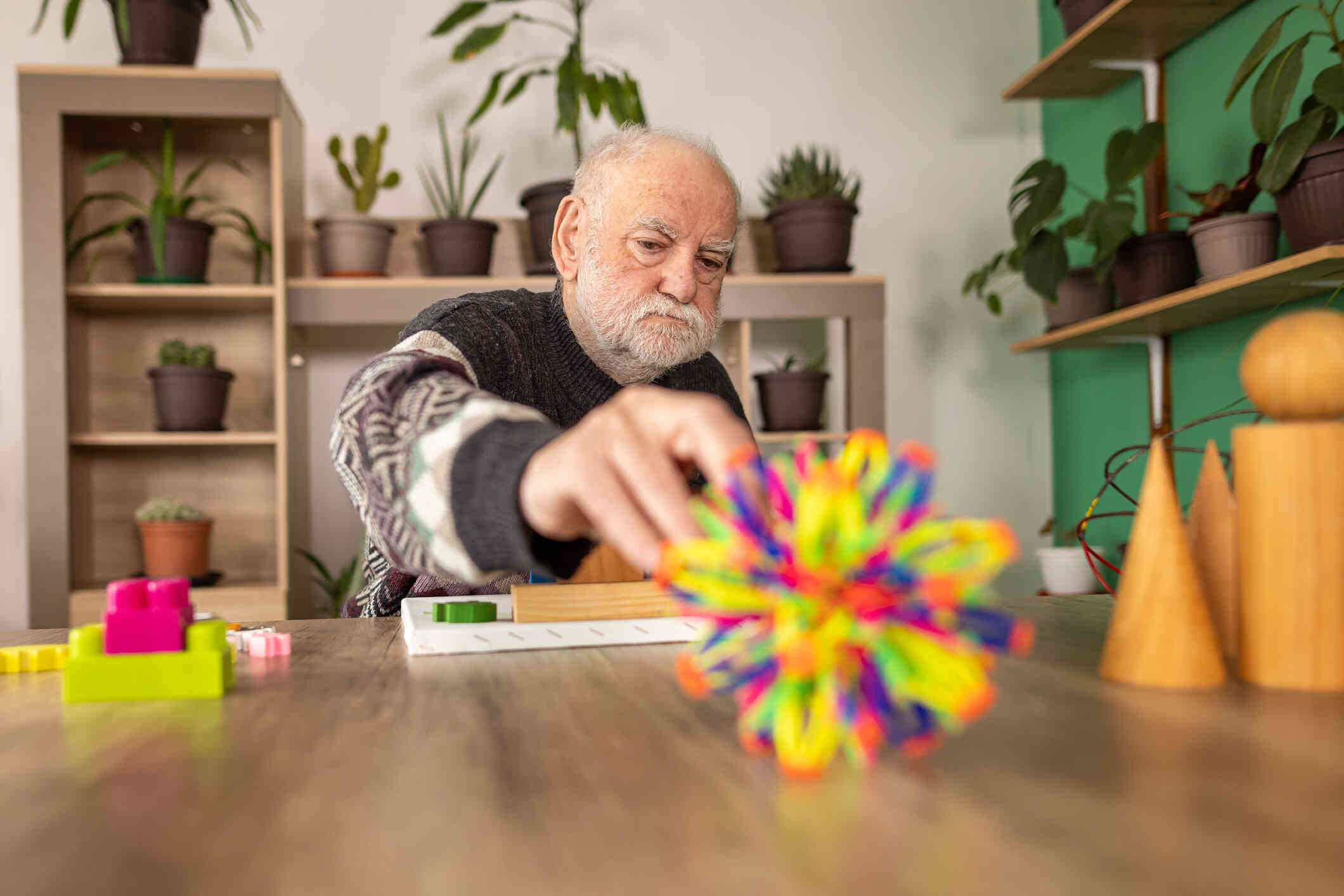 An elderly man sits at a wooden table and reaches for a toy infront of him.
