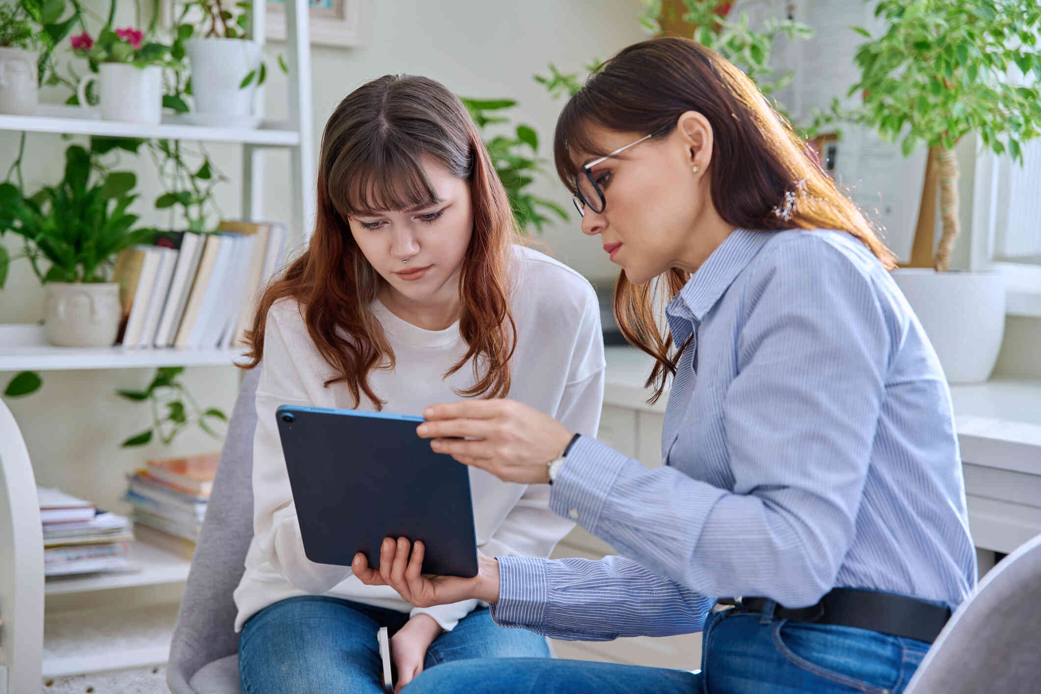 A teenage girl and a woman sit closely together, both looking at the same tablet screen the woman is holding.