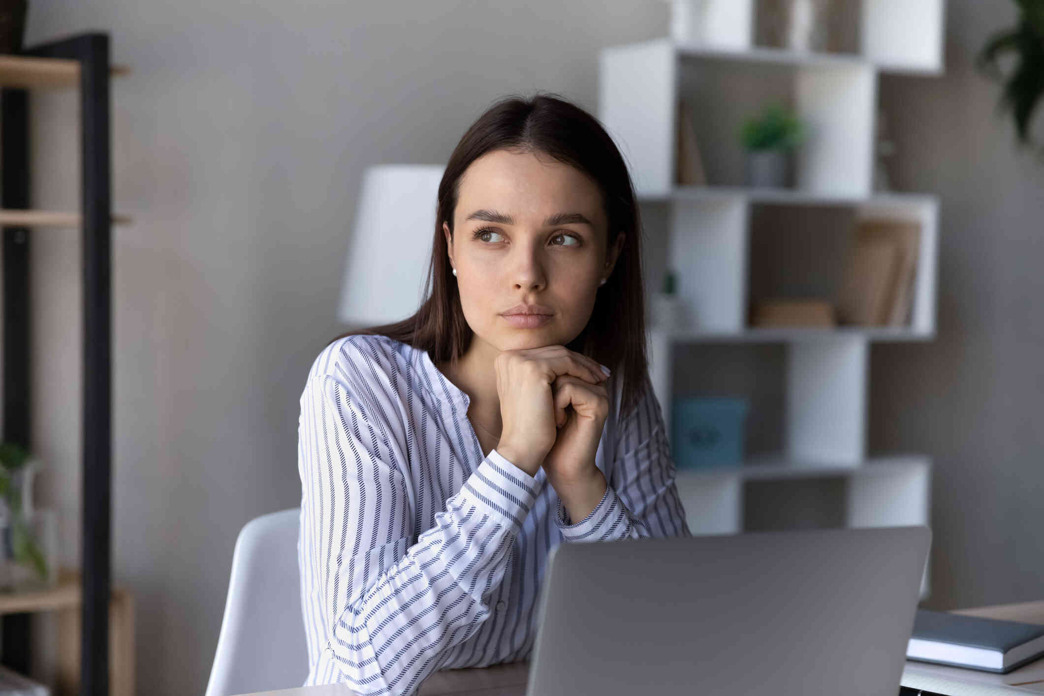 A woman in a striped shirt sits at a table with her laptop open infront of her as she gazes off with a worried expression.