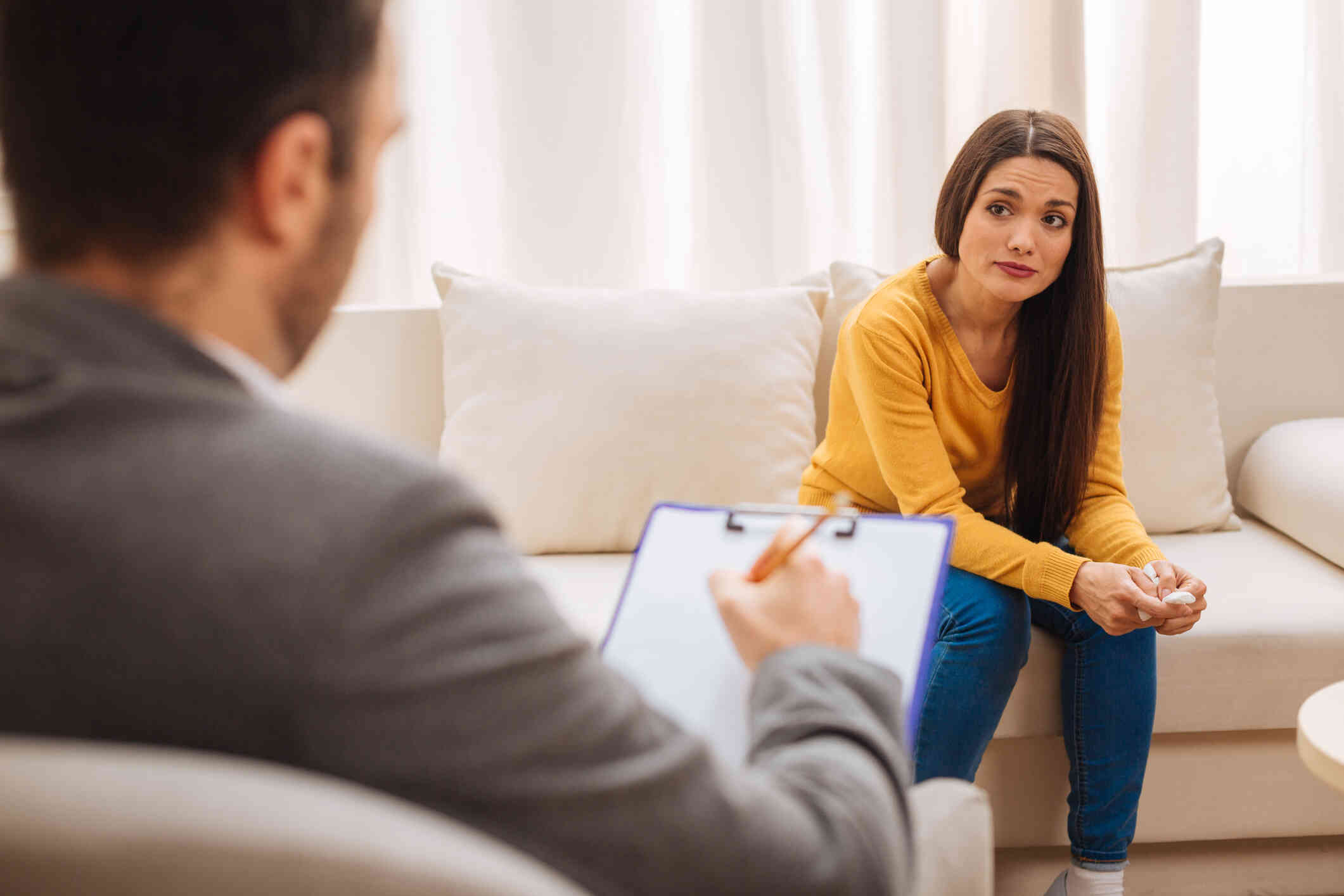 A woman in a yellow shirt leans forward sadly in her chair while looking at her male therapist during her therapy session.