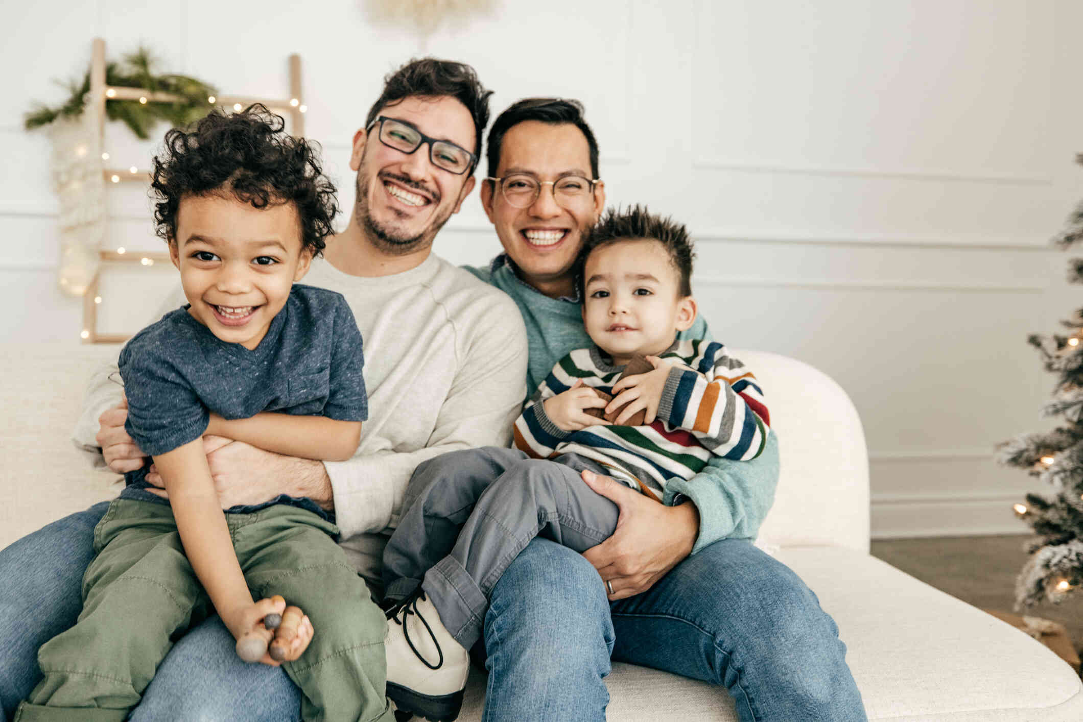 Two dads hold their young sons in their laps while smiling at the camera as they sit in their home next to each other.