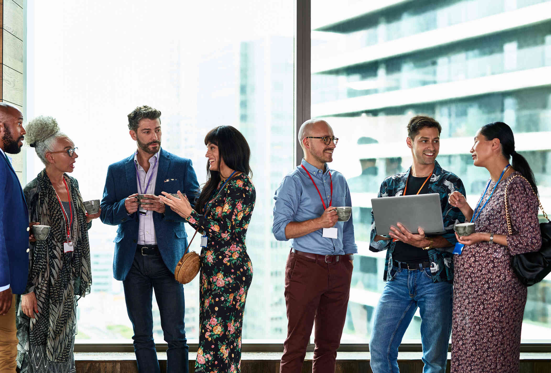 A group of men and women in business attire chat as they stand inside an office with large windows while wearing lanyards with badges attached.