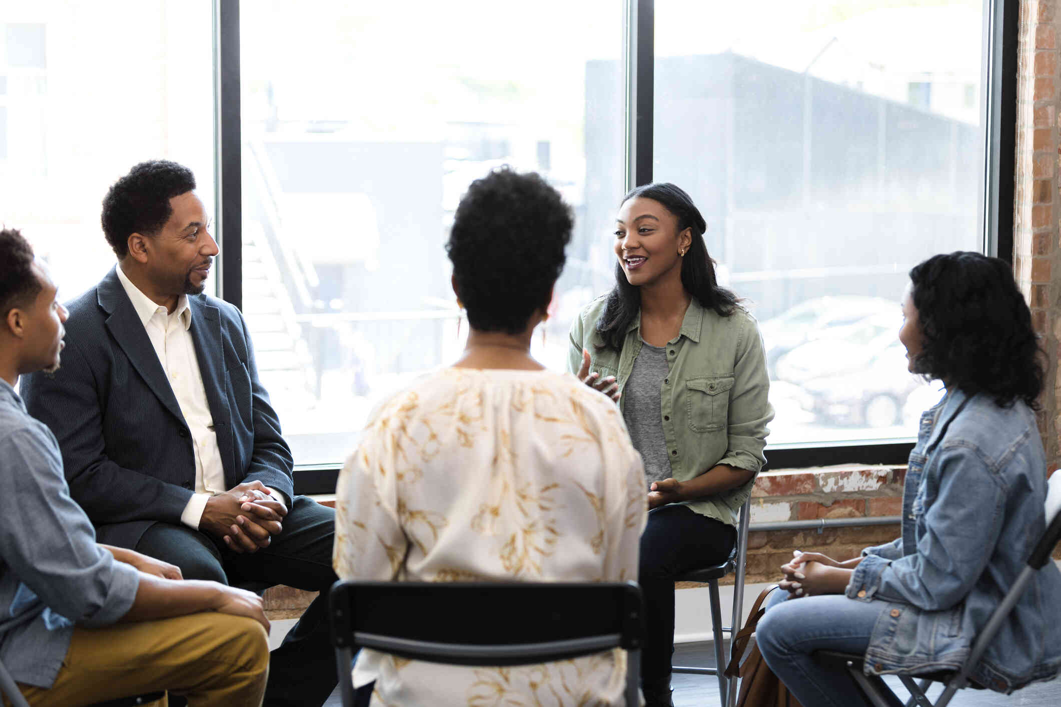 A group of adults sit in chairs in a circle with a male therapist during a grief group counseling session.