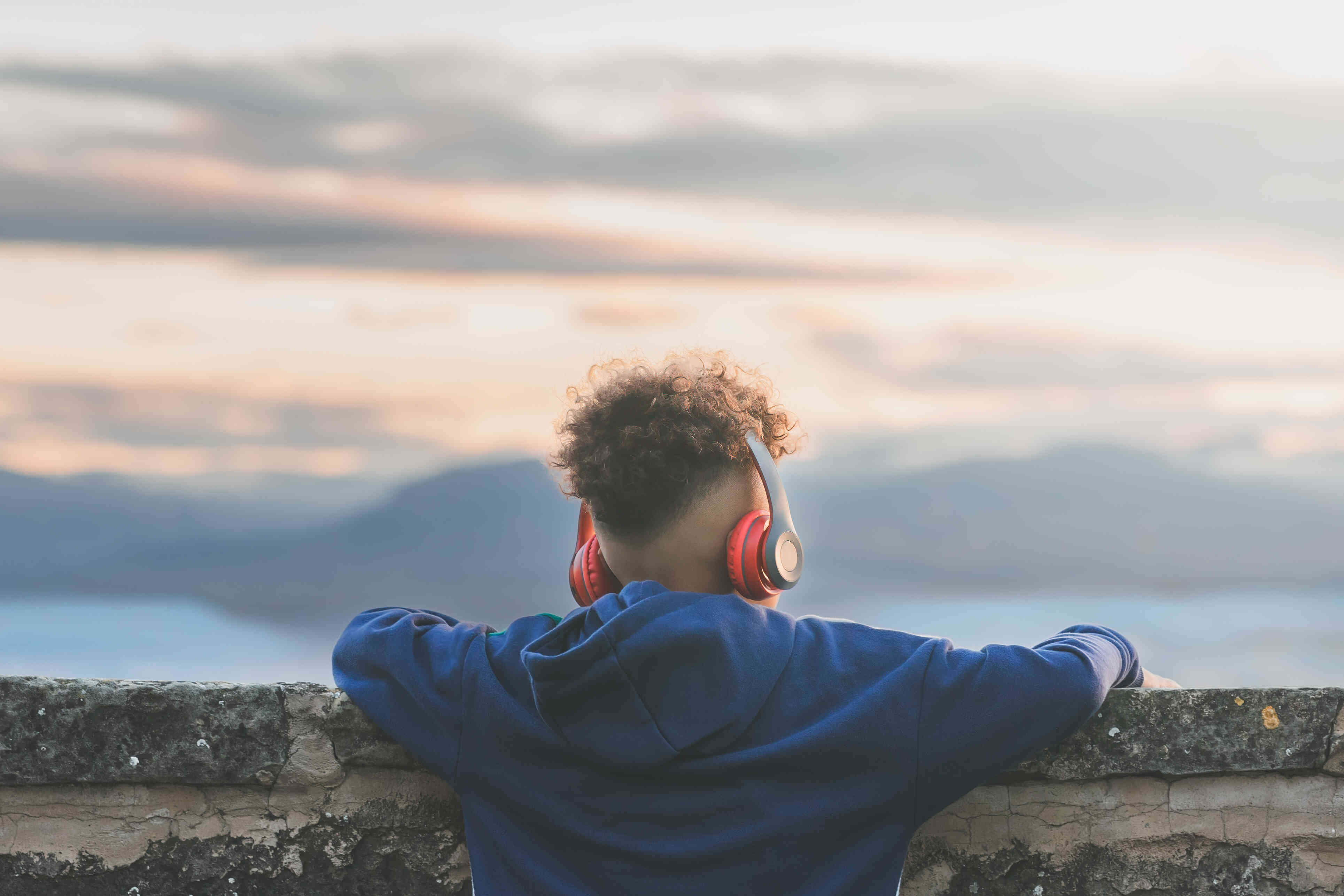 A man iwth over the ear headphones wearing a blue hoodie stands near a stone wall and watches the sunset with his back to the camera.