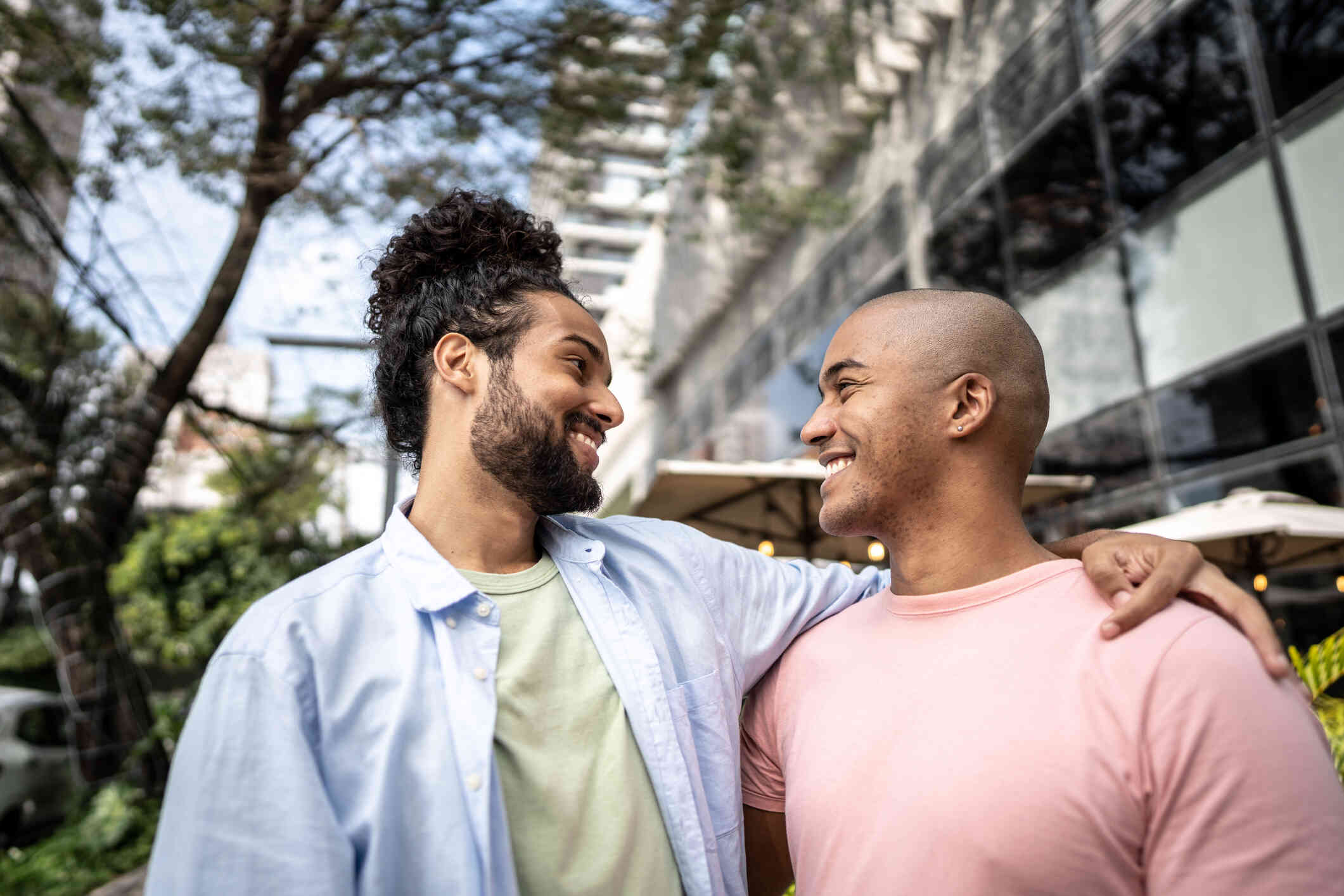 A close up of a male couple wrapping their arms around each others shoulders while smiling at one another as they stand outside on a sunny day.