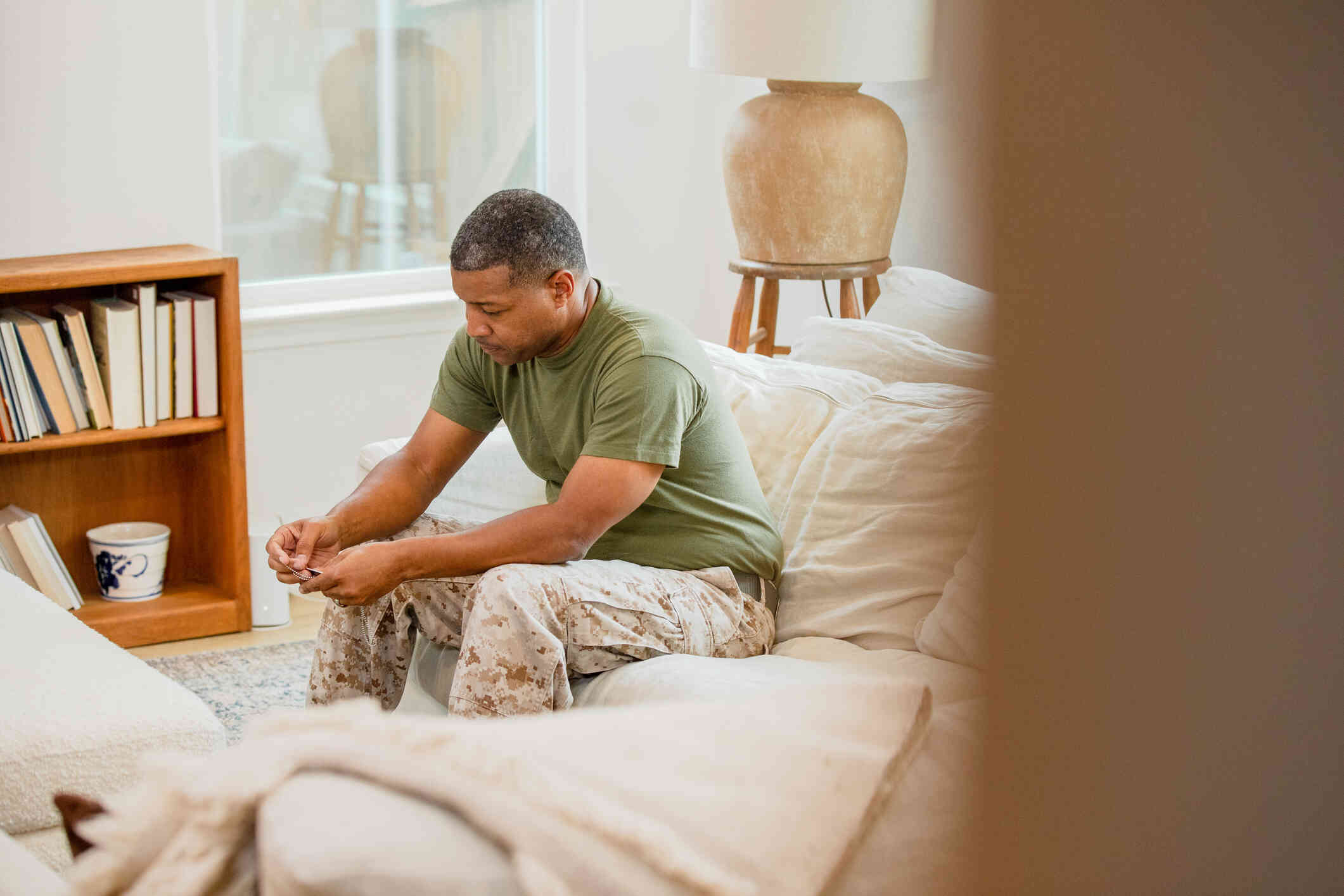 A middle aged man in an army uniform sits sadly hunched over on the couch with his head lowered.