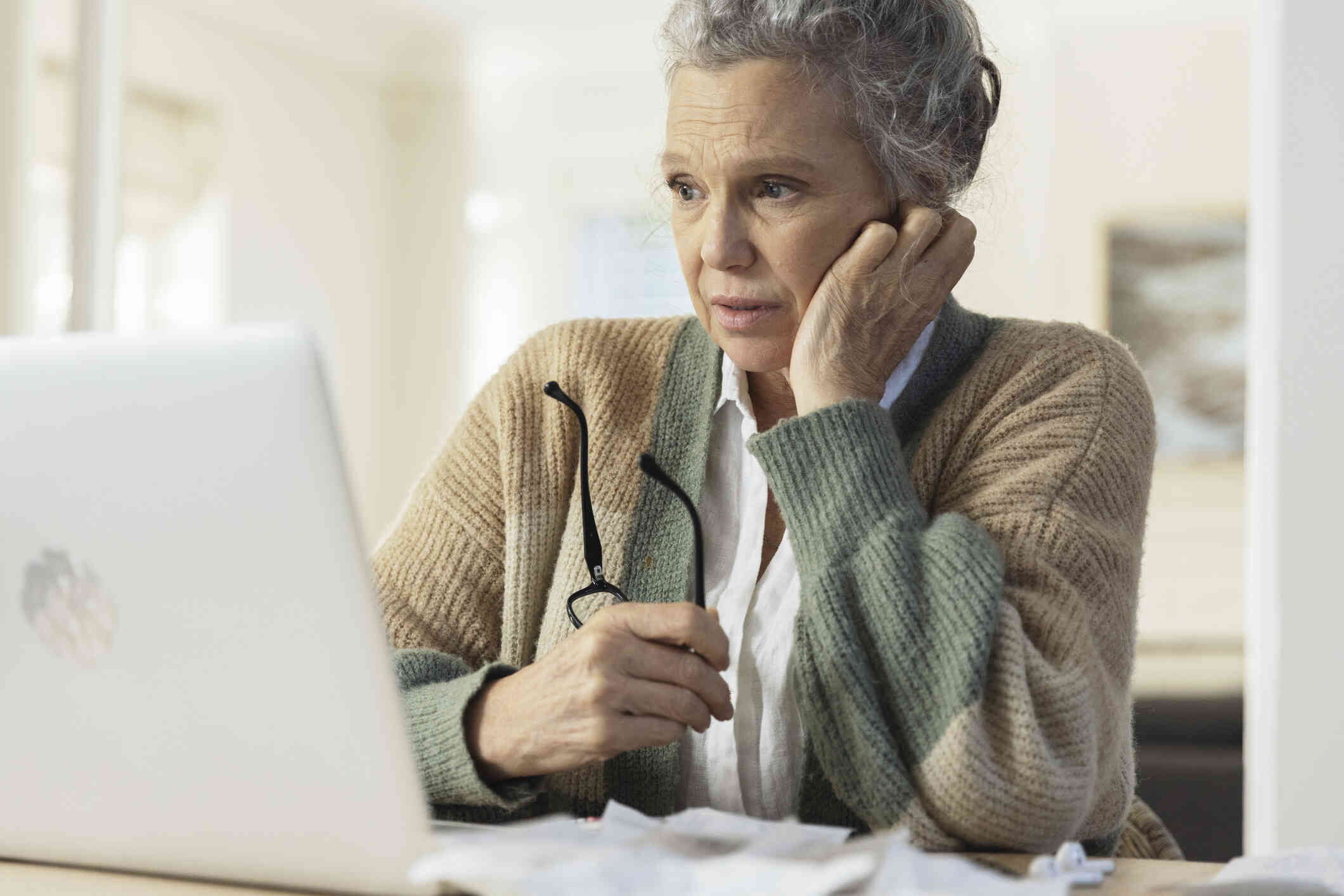 An elderly woman in a knit sweater sits at a table with her glasses in her hands as she galces off with a worried expression.